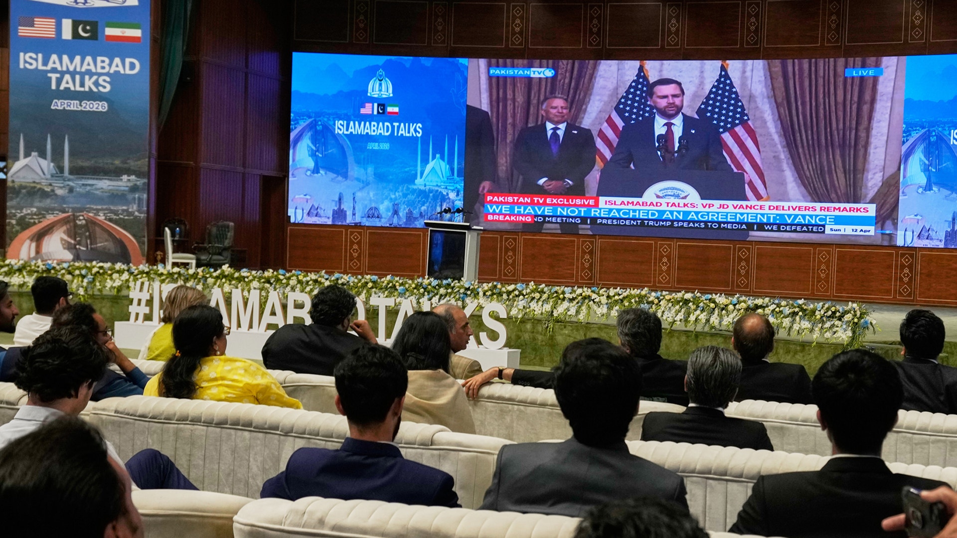 Publik sitter i en konferenssaal och tittar på en stor skärm som visar diplomater och amerikanska flaggor under ett möte i Islamabad.