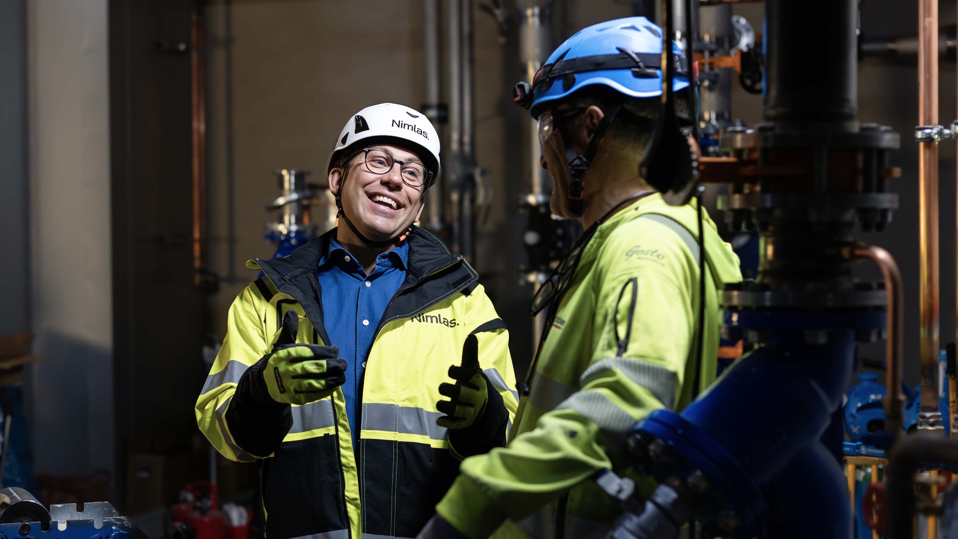 Två arbetare i säkerhetsutrustning diskuterar på en industriell arbetsplats. De bär reflekterande västar och hjälmar för säkerhet.