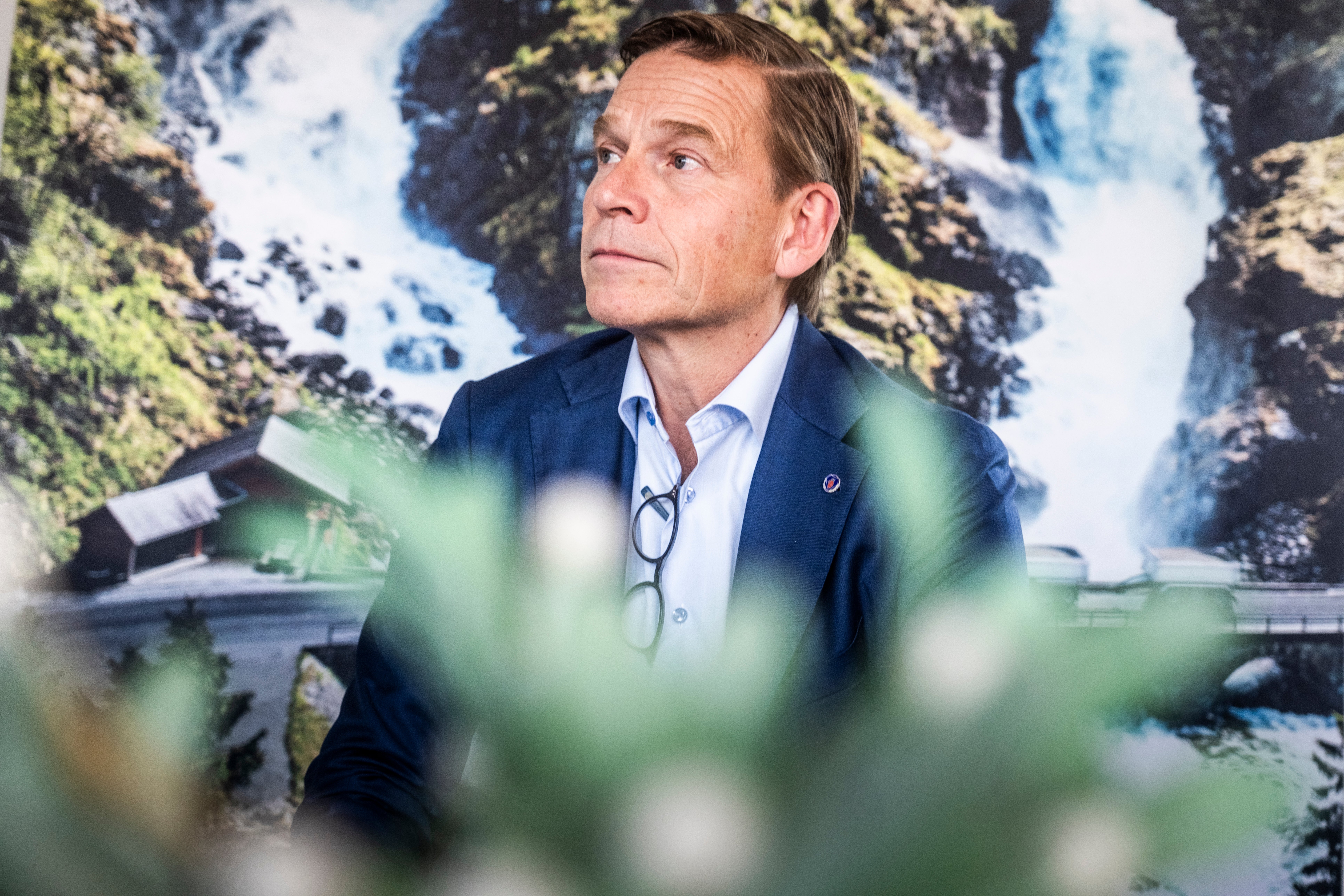 En man i mörkblå kavaj sitter framför en bakgrund med ett vattenfalls landskap. Grön växtlighet är suddig i förgrunden.
