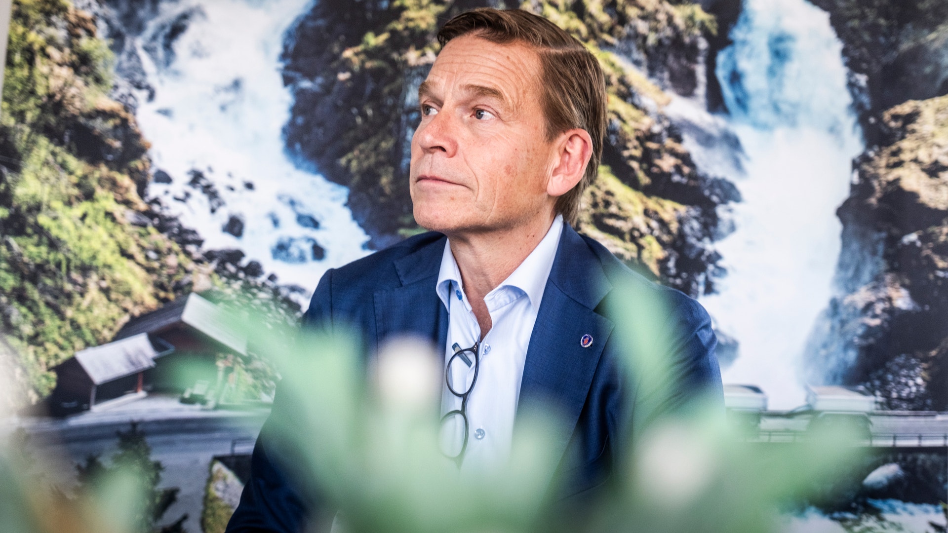 En man i mörkblå kavaj sitter framför en bakgrund med ett vattenfalls landskap. Grön växtlighet är suddig i förgrunden.