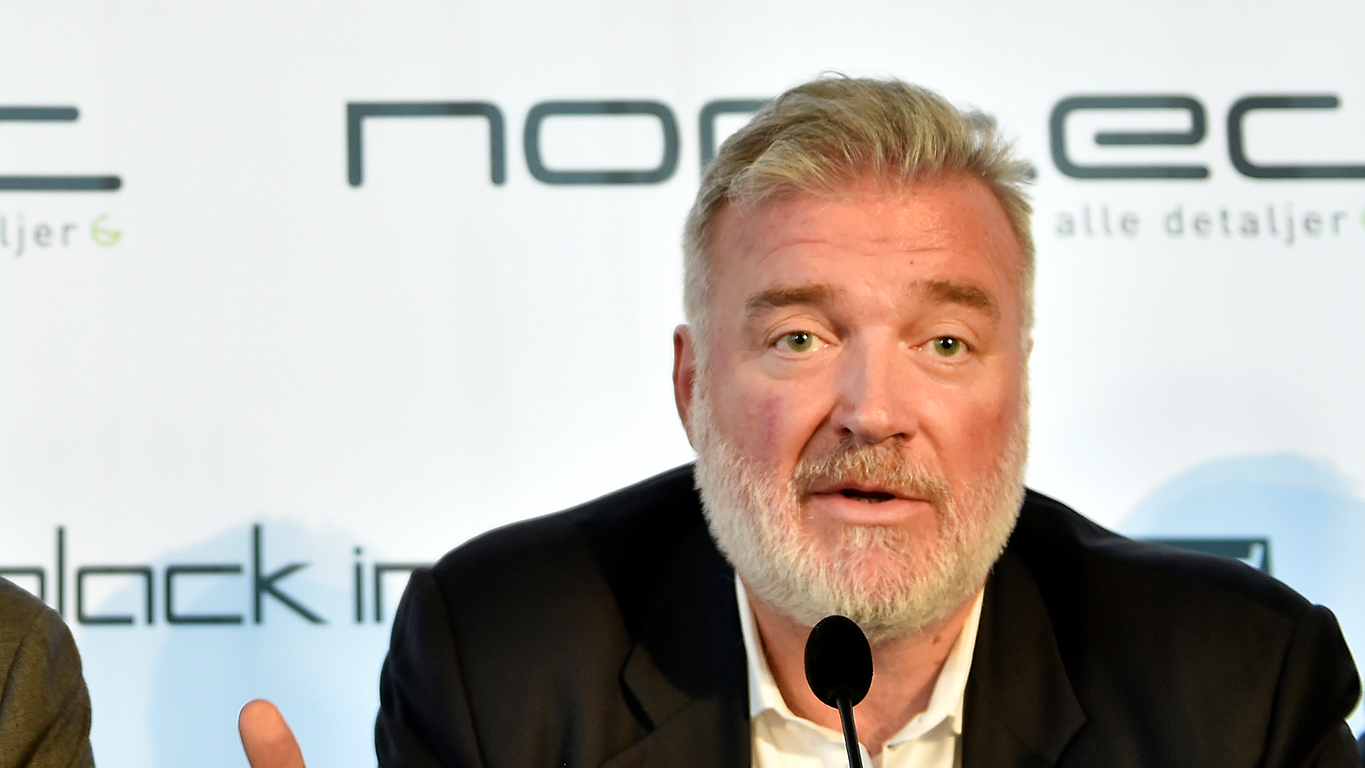 Porträttfoto av en man med grått hår och skägg som talar vid ett konferenspanel