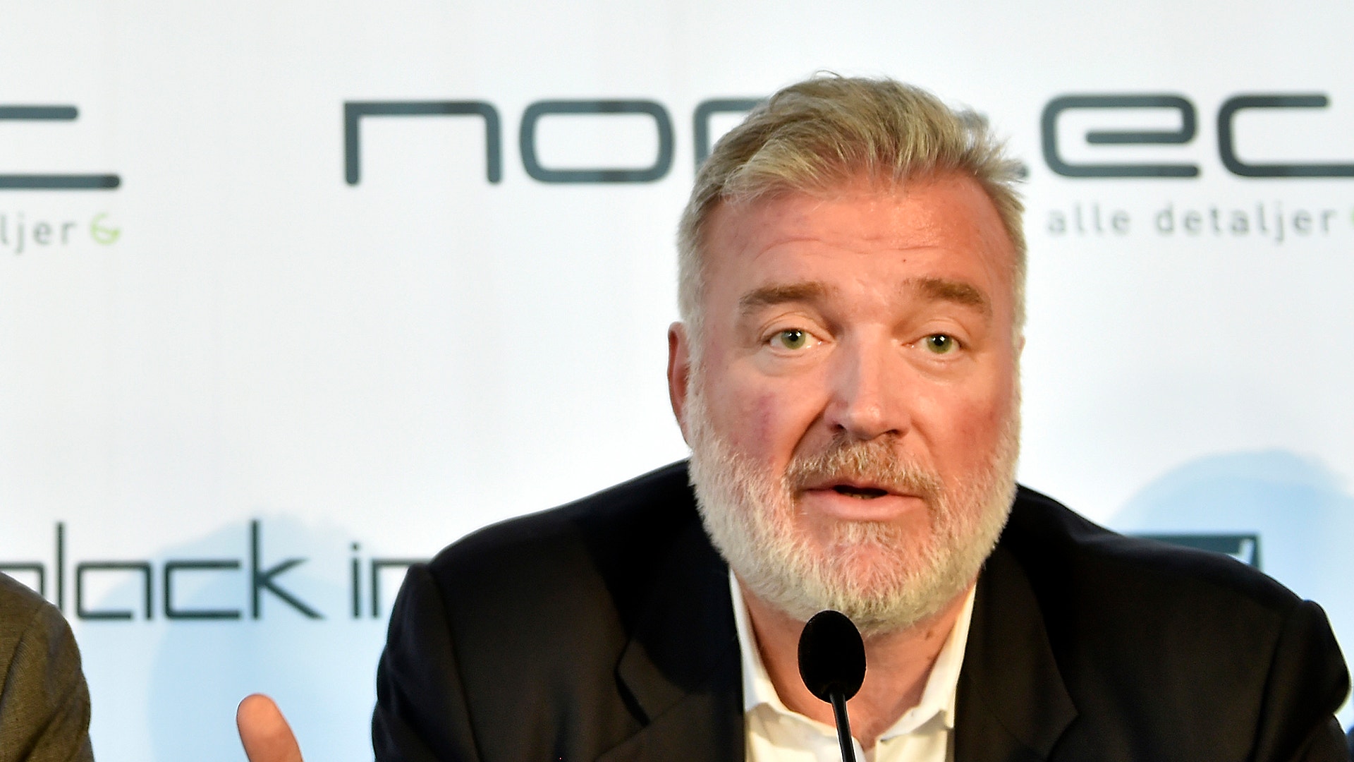 Porträttfoto av en man med grått hår och skägg som talar vid ett konferenspanel