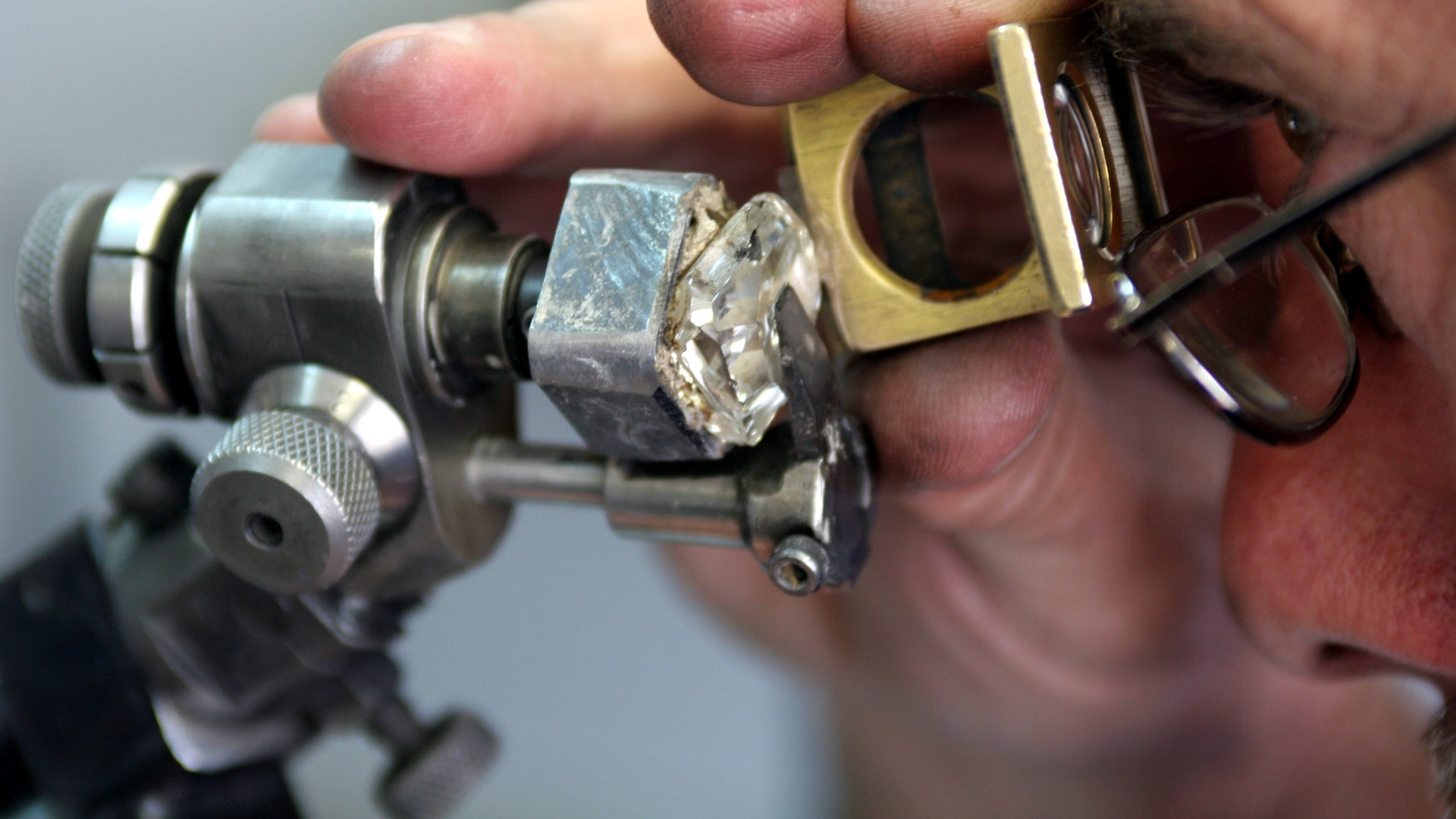 En man med glasögon granskar en oslipad diamant med ett förstoringsglas och ett mätinstrument.