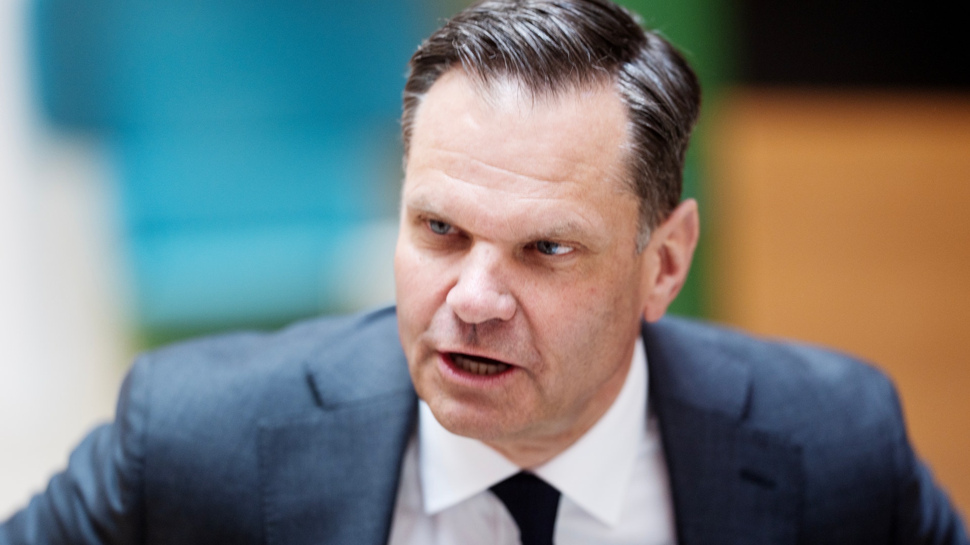 Porträtt av en man i mörkblå kostym och svart slips som talar eller presenterar något.