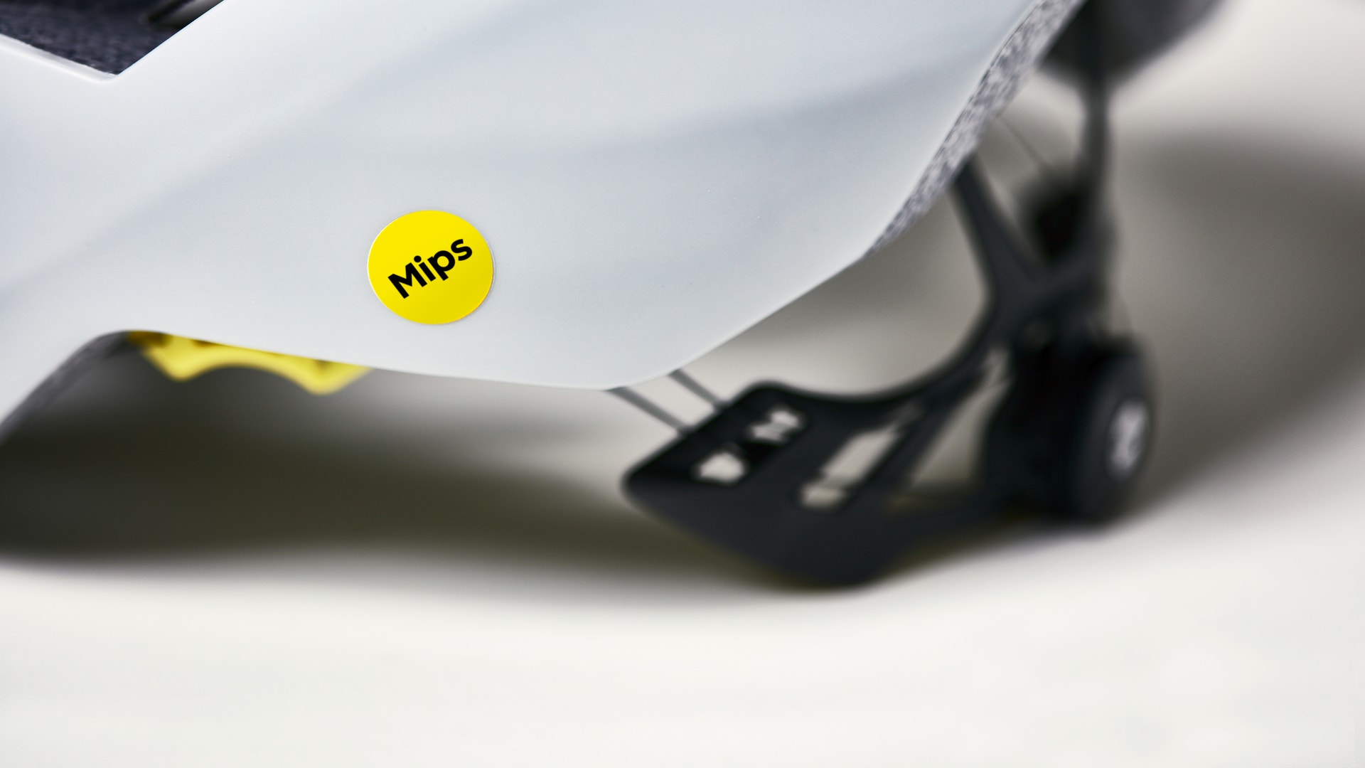 Närbildsfotografi av en vit cykelhjälm med Mips-skyddssystem. Det gula Mips-logotypen är synlig på hjälmen.