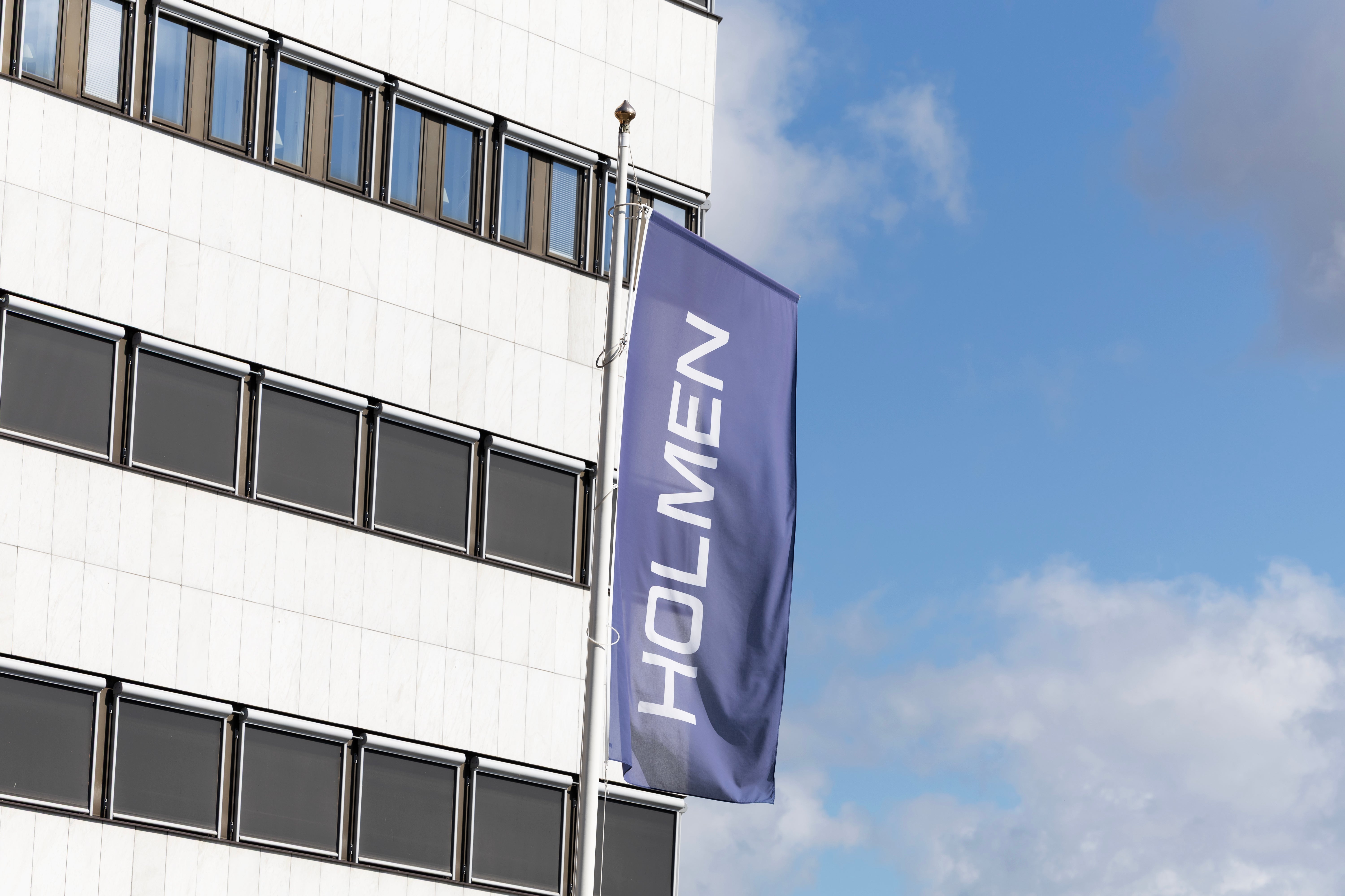 Vit modern byggnad med ett blått banner med texten HOLMEN hängande på fasaden. Blå himmel med moln i bakgrunden.