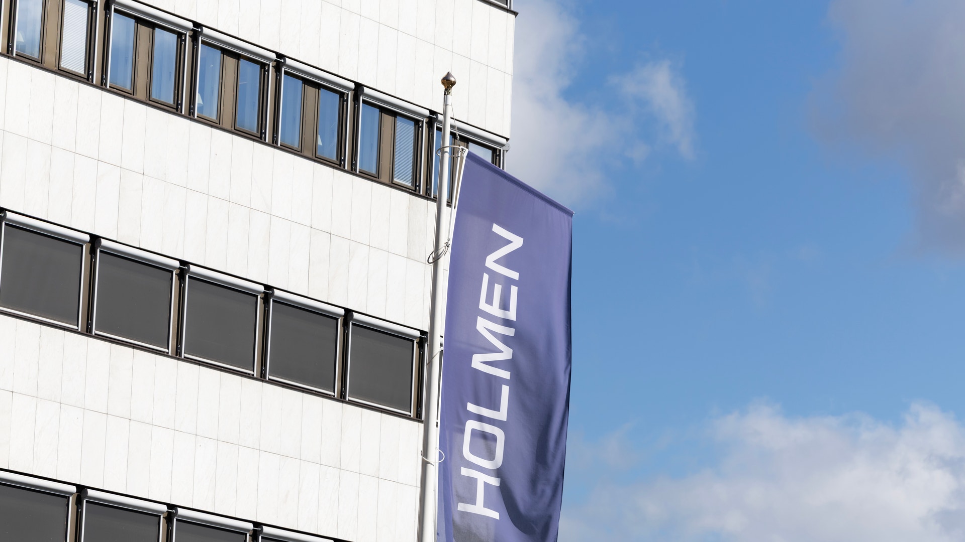Vit modern byggnad med ett blått banner med texten HOLMEN hängande på fasaden. Blå himmel med moln i bakgrunden.