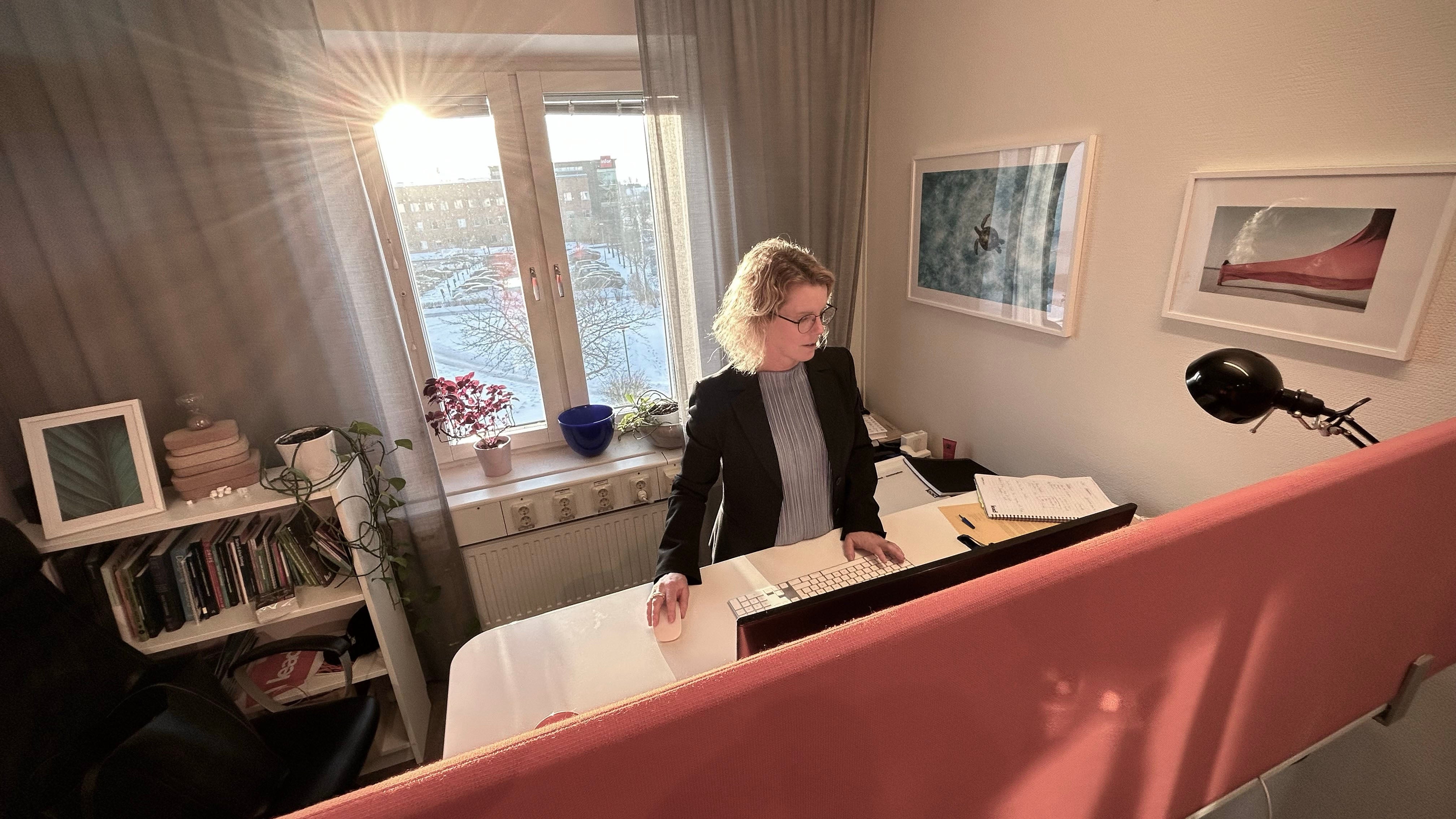 Person i svart kavaj sitter vid skrivbord i modernt kontor med utsikt över stad genom stort fönster. Rummet är inrett med böcker, växter och konstverk.