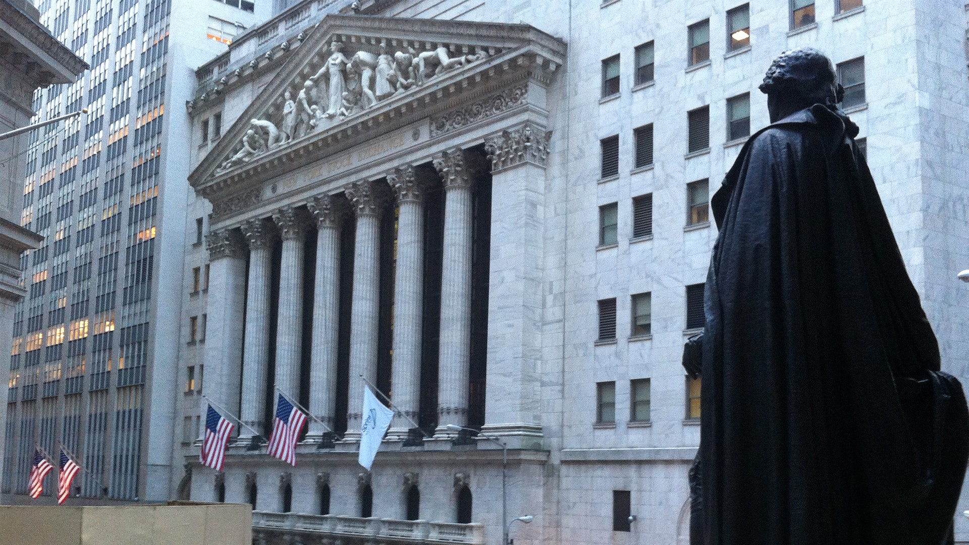 Statyn framför New York Stock Exchange-byggnaden med dess klassiska vita kolonner och amerikanska flaggor.