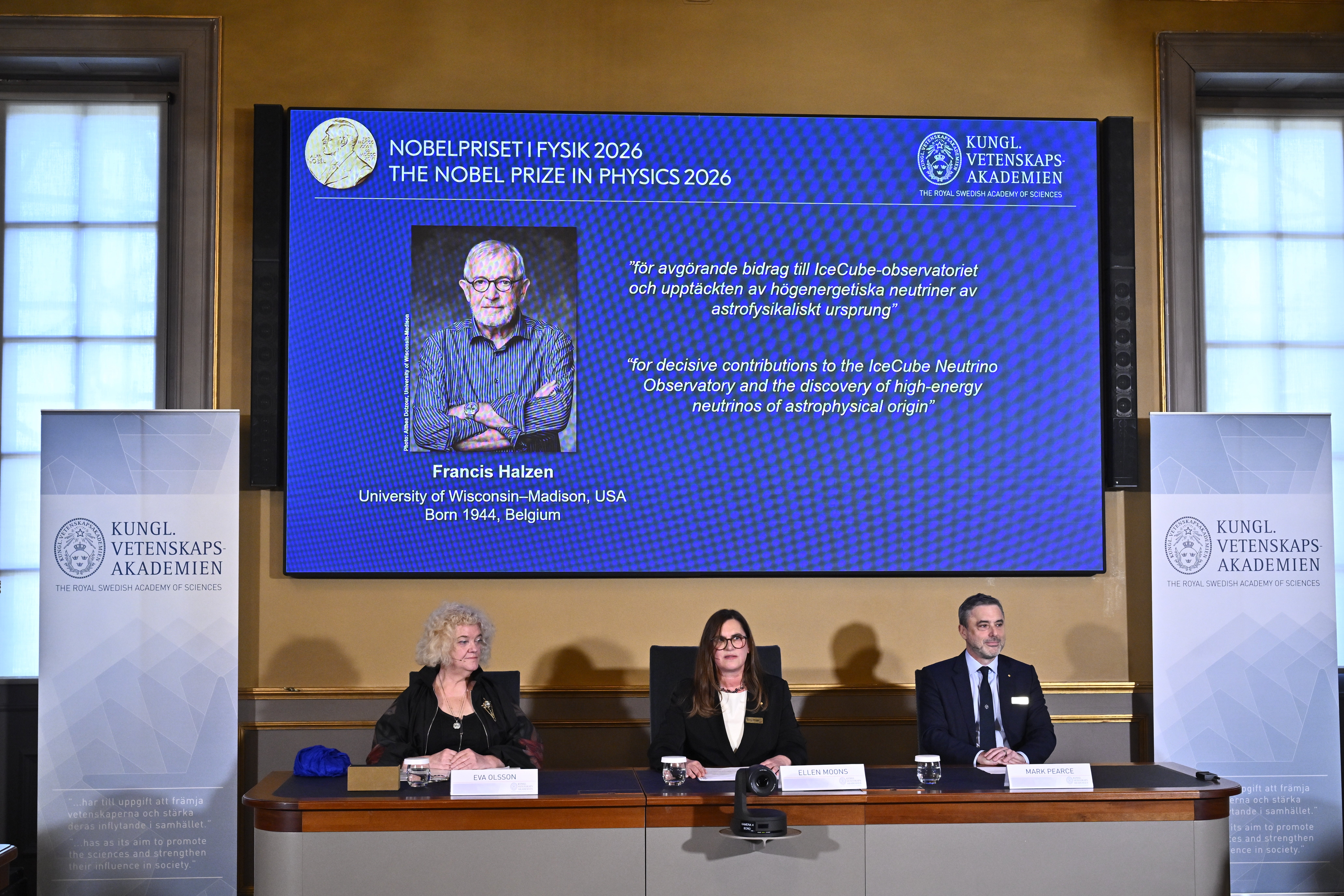 Tre personer sitter vid ett bord under en pressträff. Bakom dem visar en stor skärm Nobelpriset i fysik 2026 med ett porträtt av pristagaren.