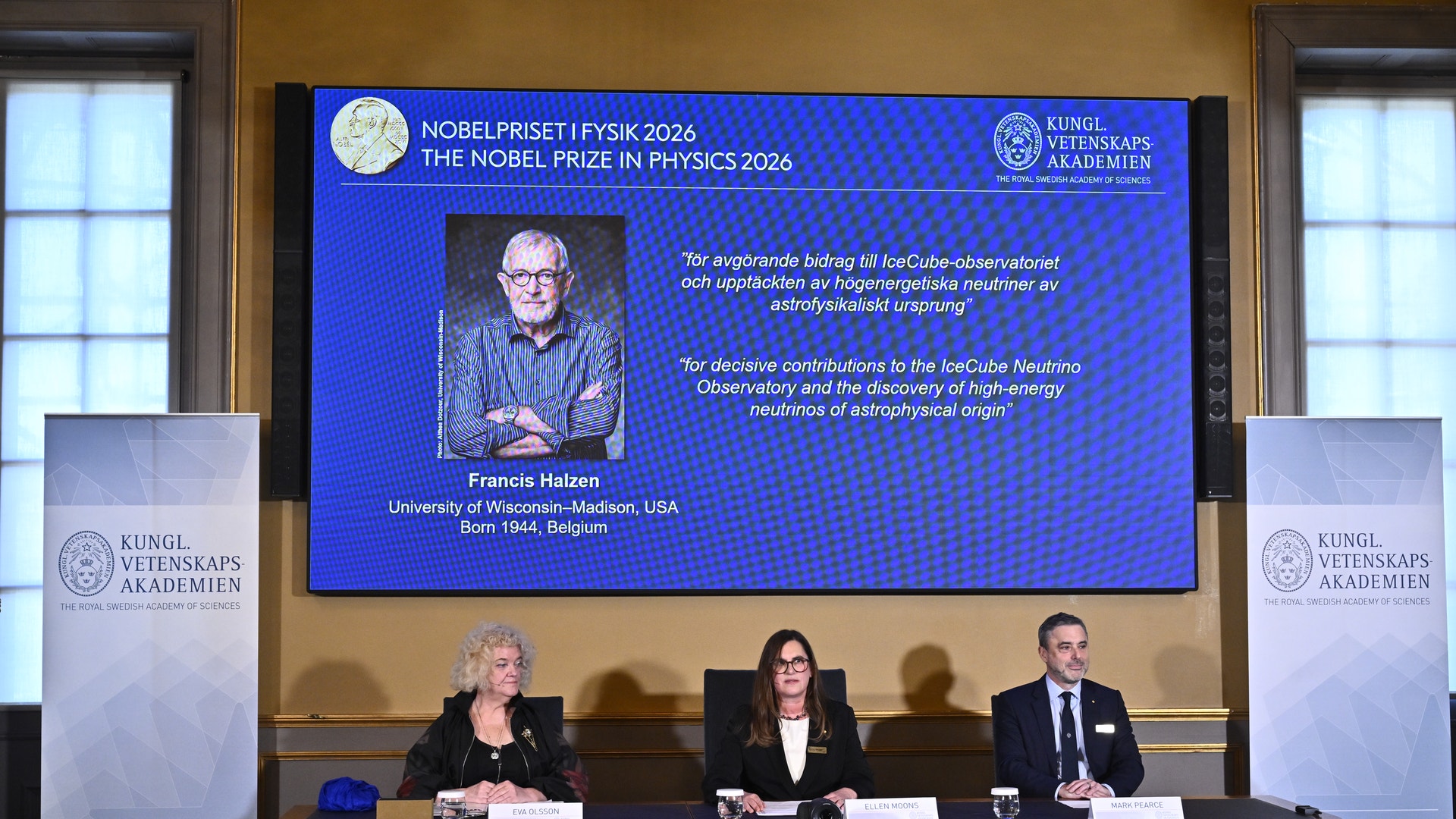 Tre personer sitter vid ett bord under en pressträff. Bakom dem visar en stor skärm Nobelpriset i fysik 2026 med ett porträtt av pristagaren.