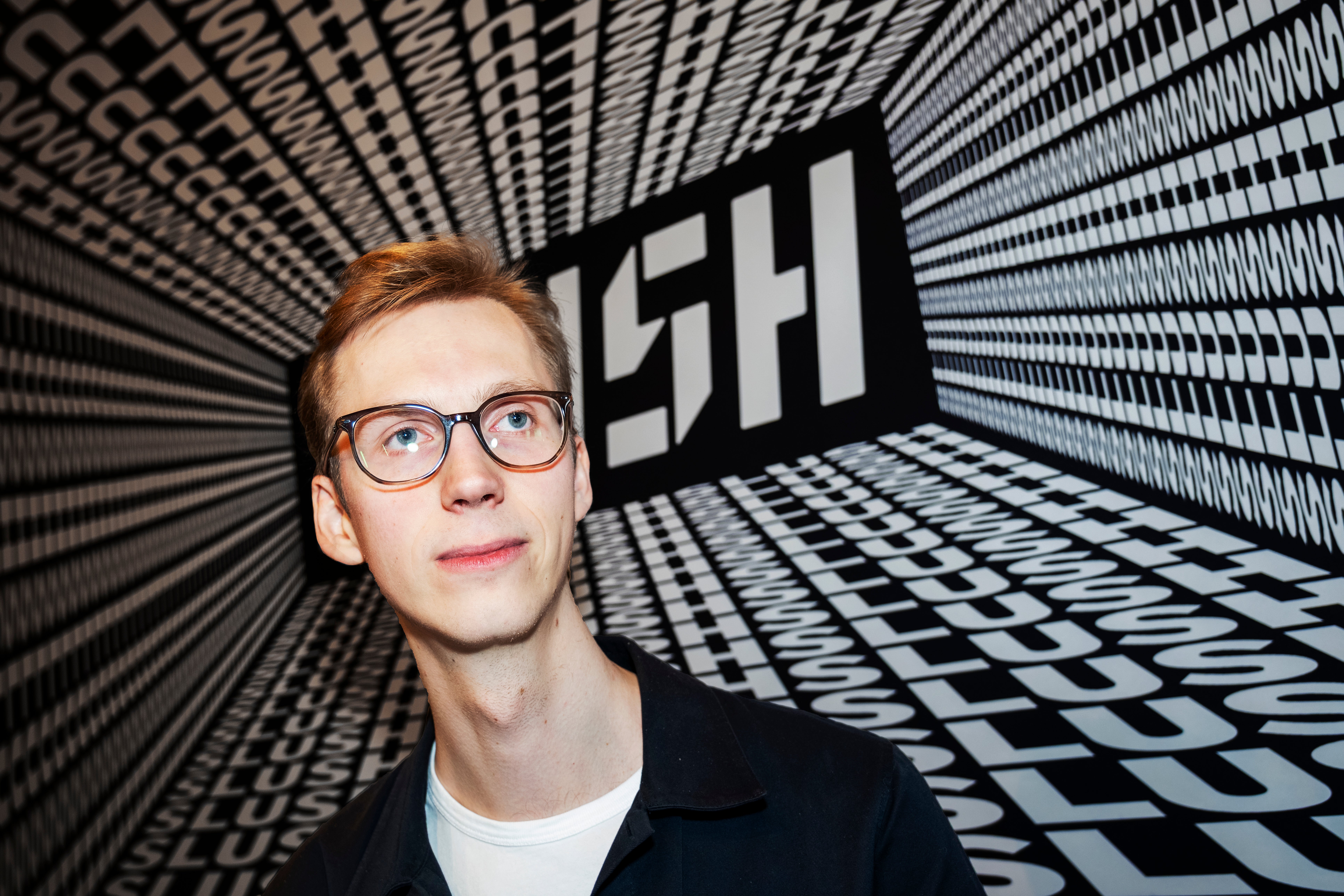 Porträtt av en ung man med rödbrunt hår och glasögon, klädd i svart jacka över vit skjorta, framför en dramatisk svart-vit geometrisk bakgrund med strålande linjer och rutmönster som skapar djup och rörelse.