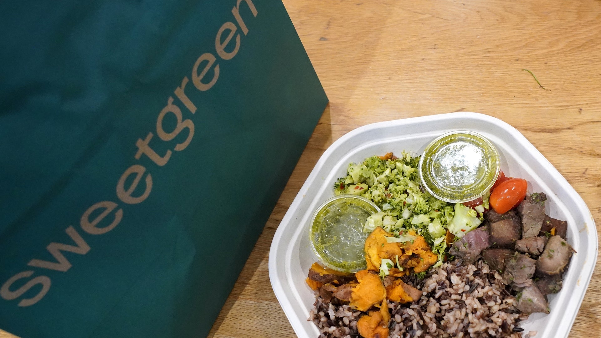 En måltidslåda från Sweetgreen med grönkålssallad, rostad sötpotatis, köttfärs och två såser i små behållare, serverad i en vit plastbehållare på ett träbord.