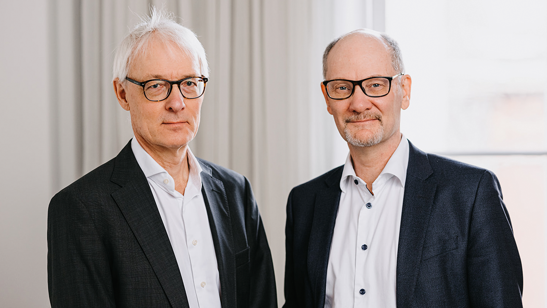 Porträtt av två män i affärskläder som står sida vid sida. Båda bär glasögon och mörka blazers över ljusa skjortor.