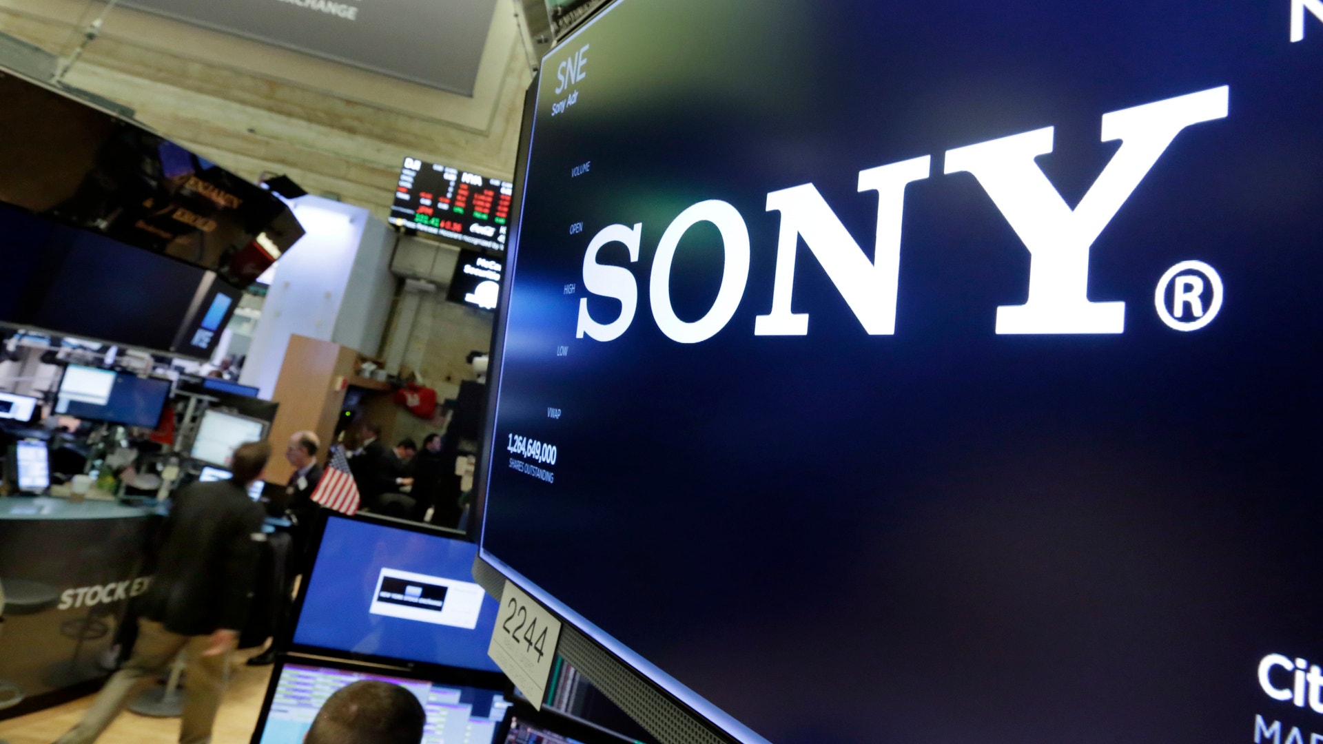 Sony-logotyp på en stor skärm på en handelsplats med arbetare och datorer i bakgrunden.
