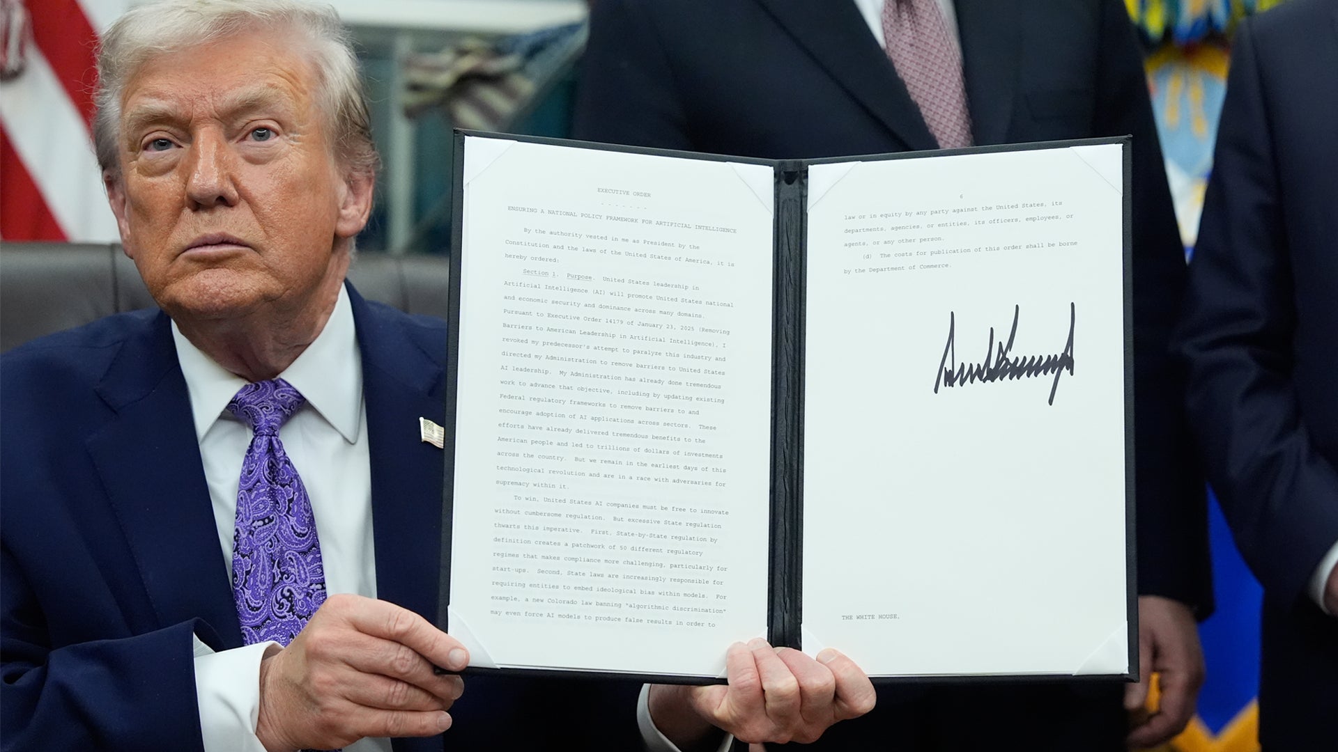 Man i formell kostym håller upp ett signerat dokument framför en amerikansk flagga.