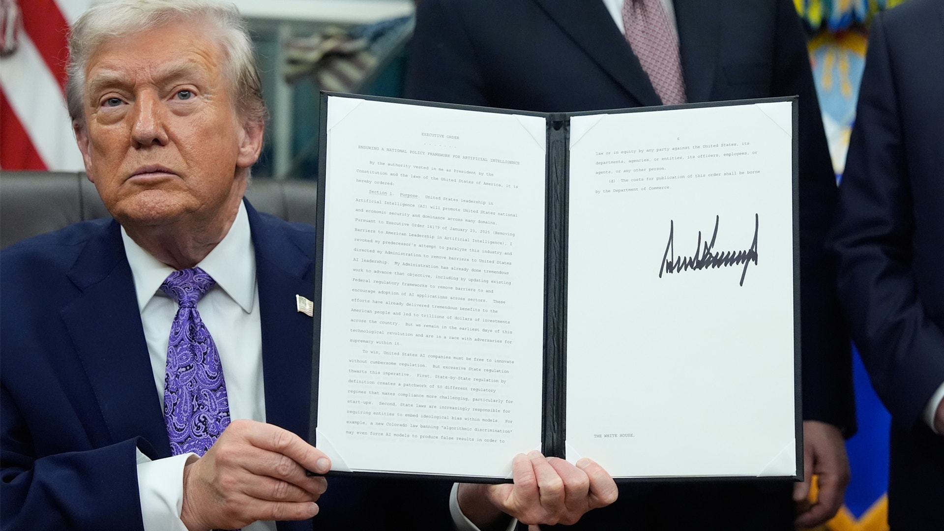 Man i formell kostym håller upp ett signerat dokument framför en amerikansk flagga.