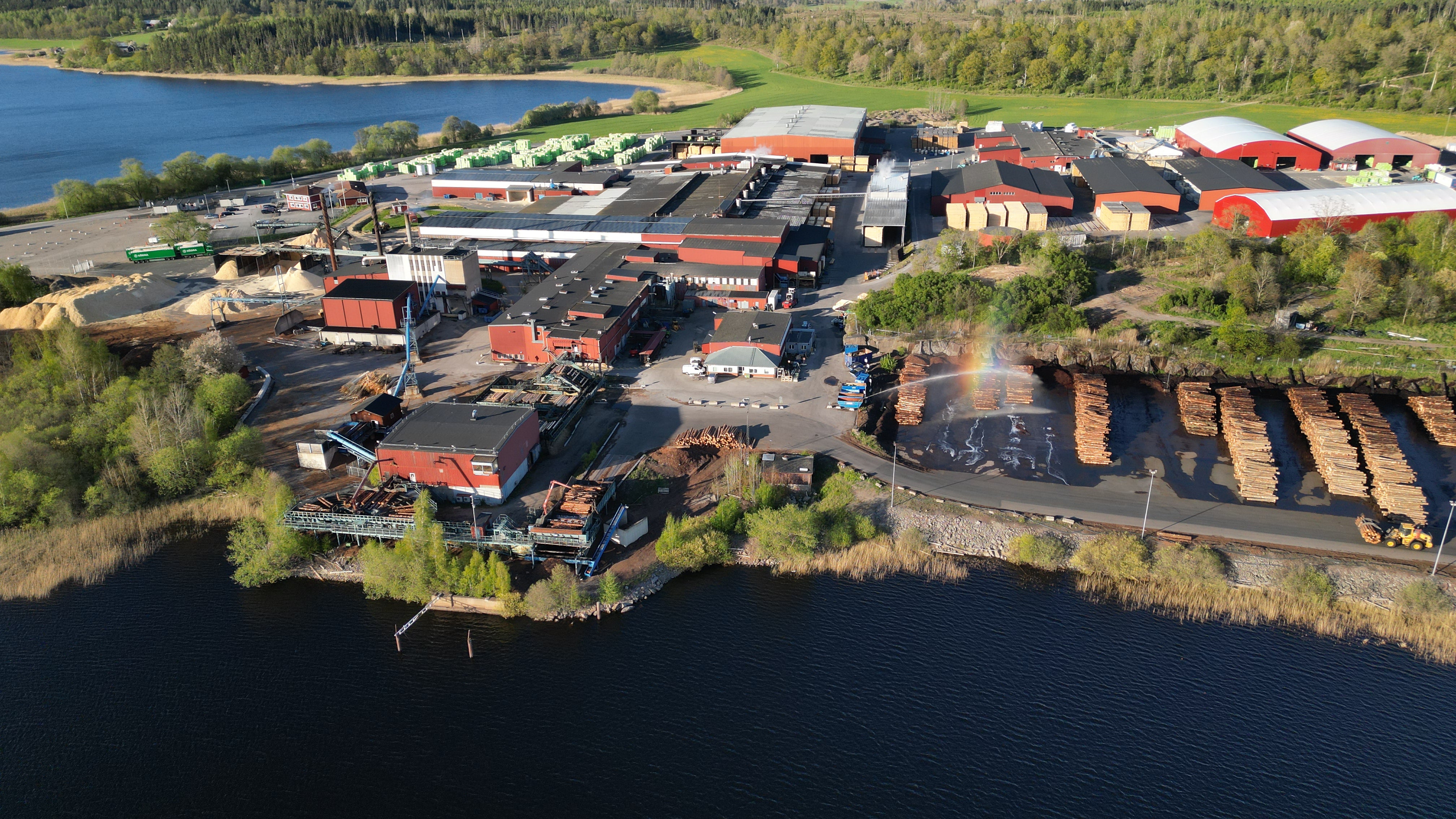 Flygfoto av en industriell anläggning med flera byggnader och röda tak vid en sjö, omgiven av skog och gröna marker.