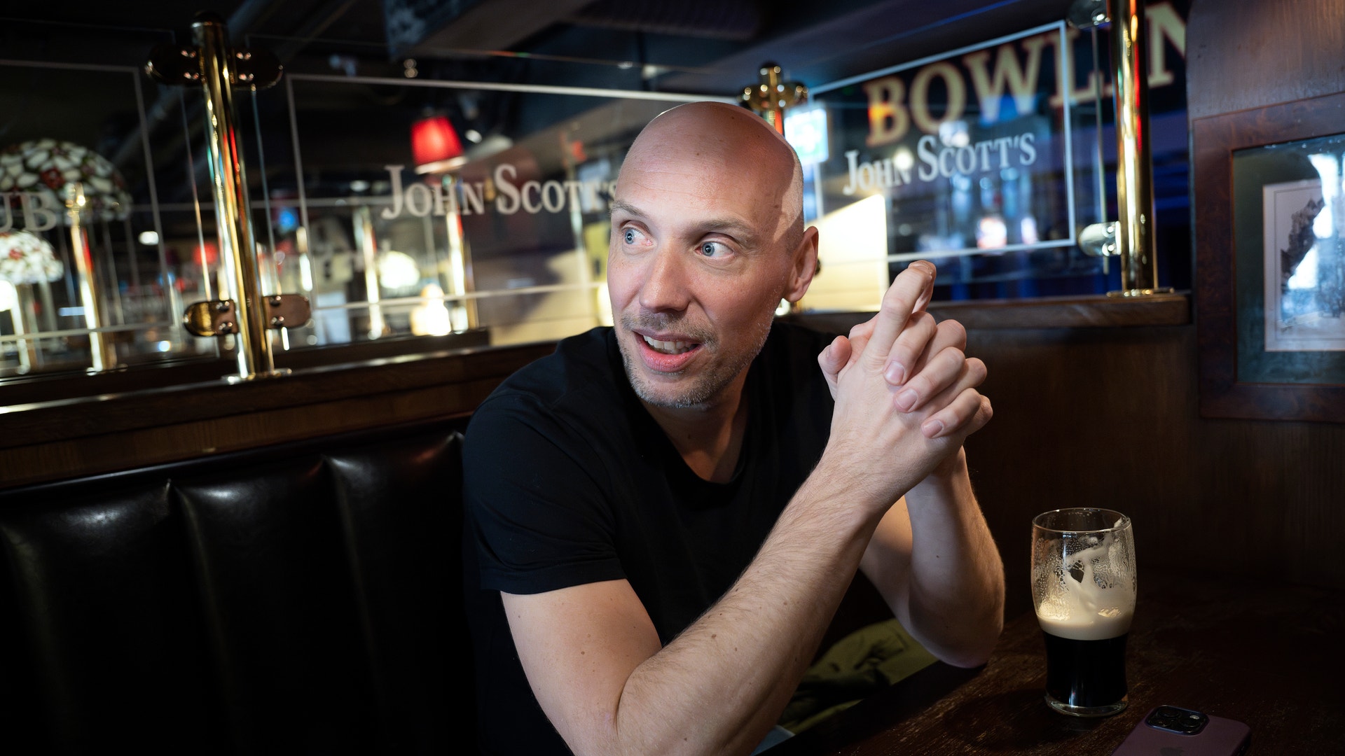 En man i svart skjorta sitter vid en bardisk med ett ölglas framför sig.