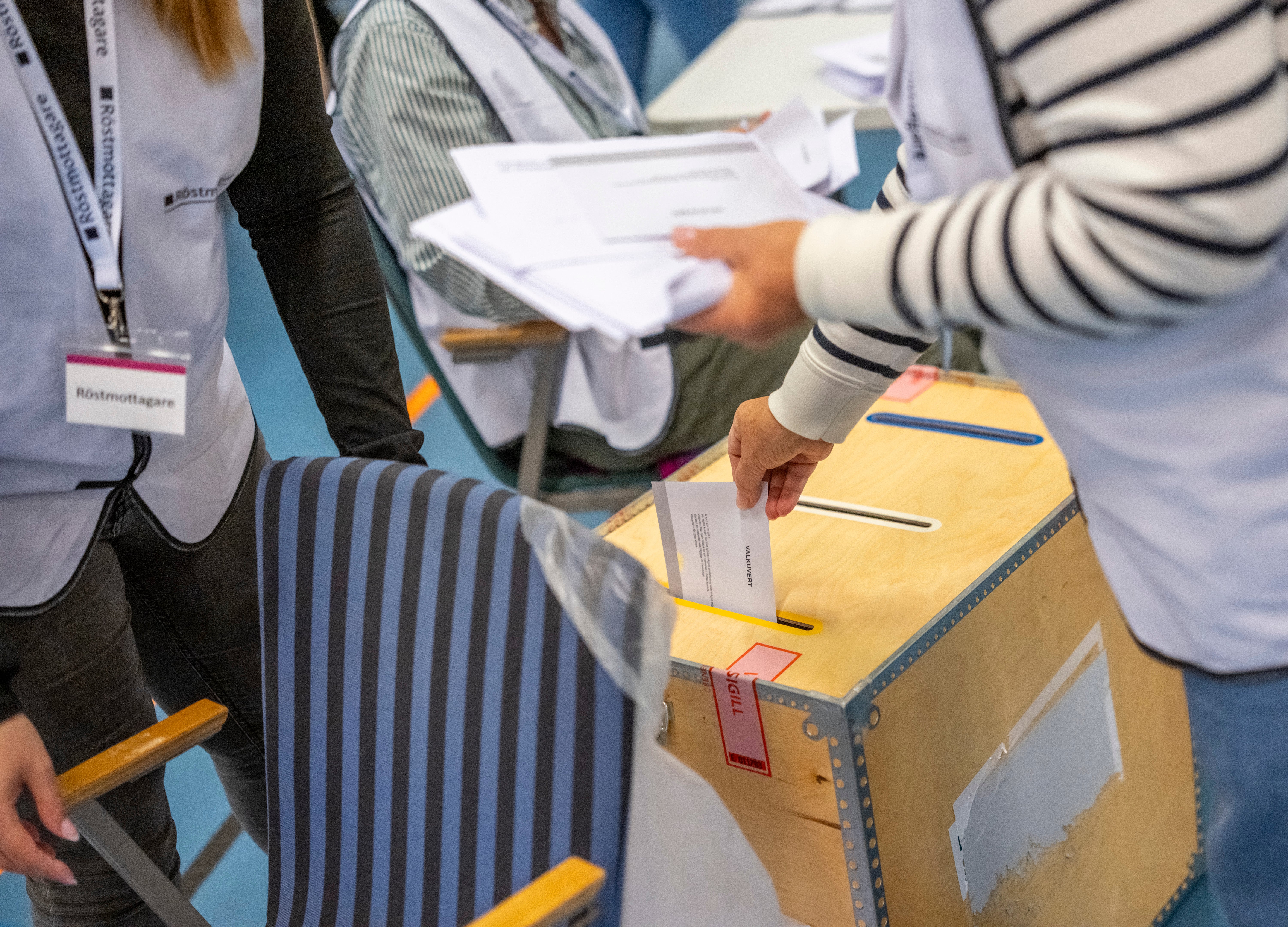 En person lägger sin röst i en valurna medan en röstmottagare håller i valsedlar i bakgrunden.