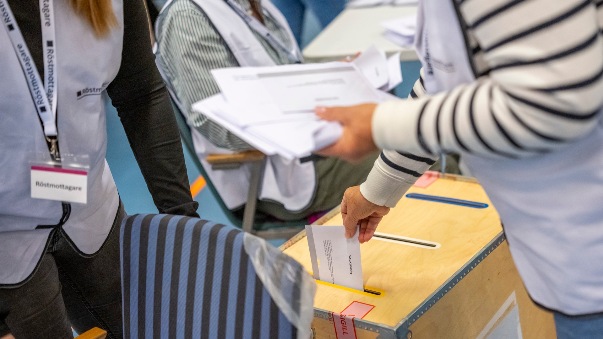 En person lägger sin röst i en valurna medan en röstmottagare håller i valsedlar i bakgrunden.