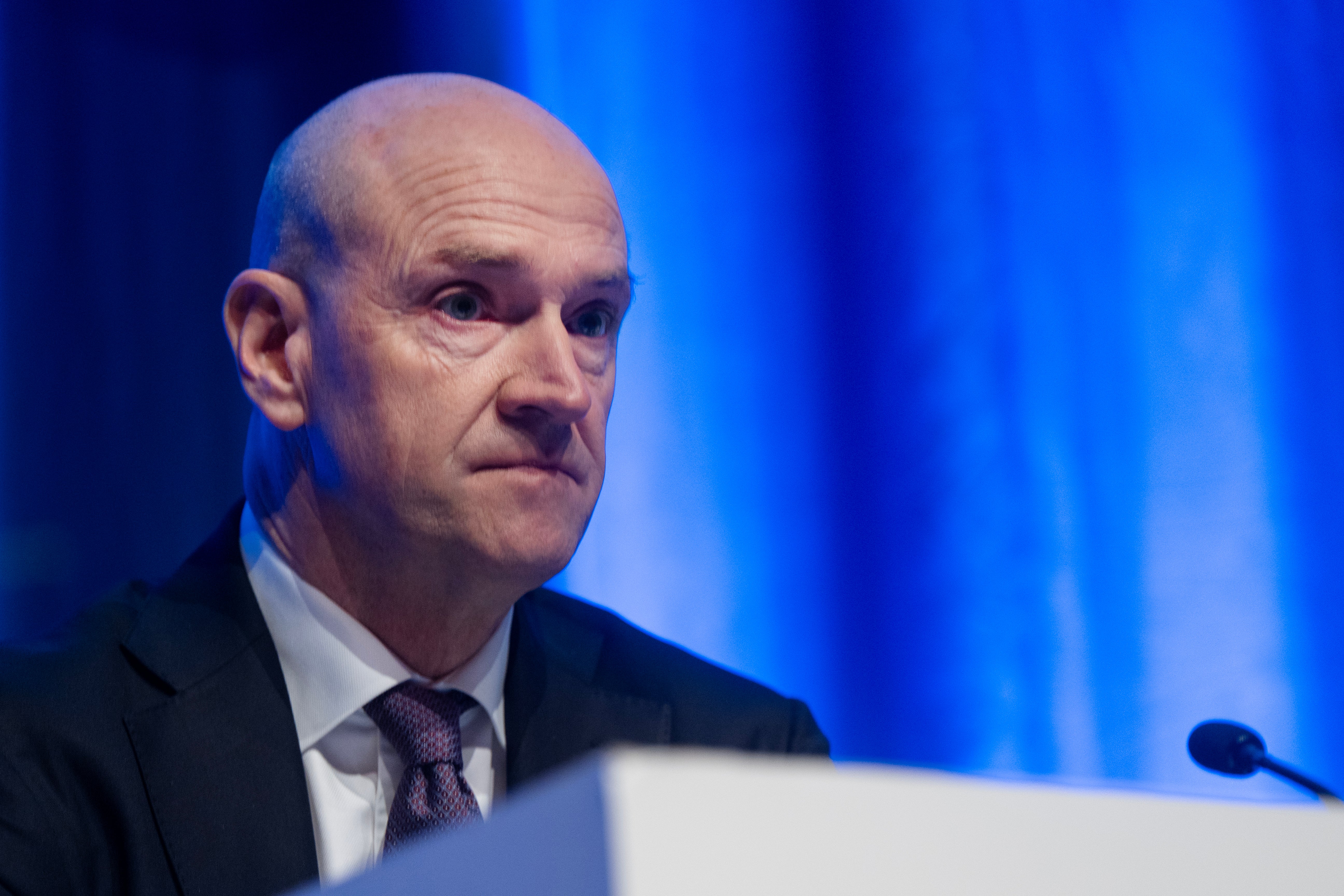 En man i kostym och slips talar vid ett podium mot en blå bakgrund.