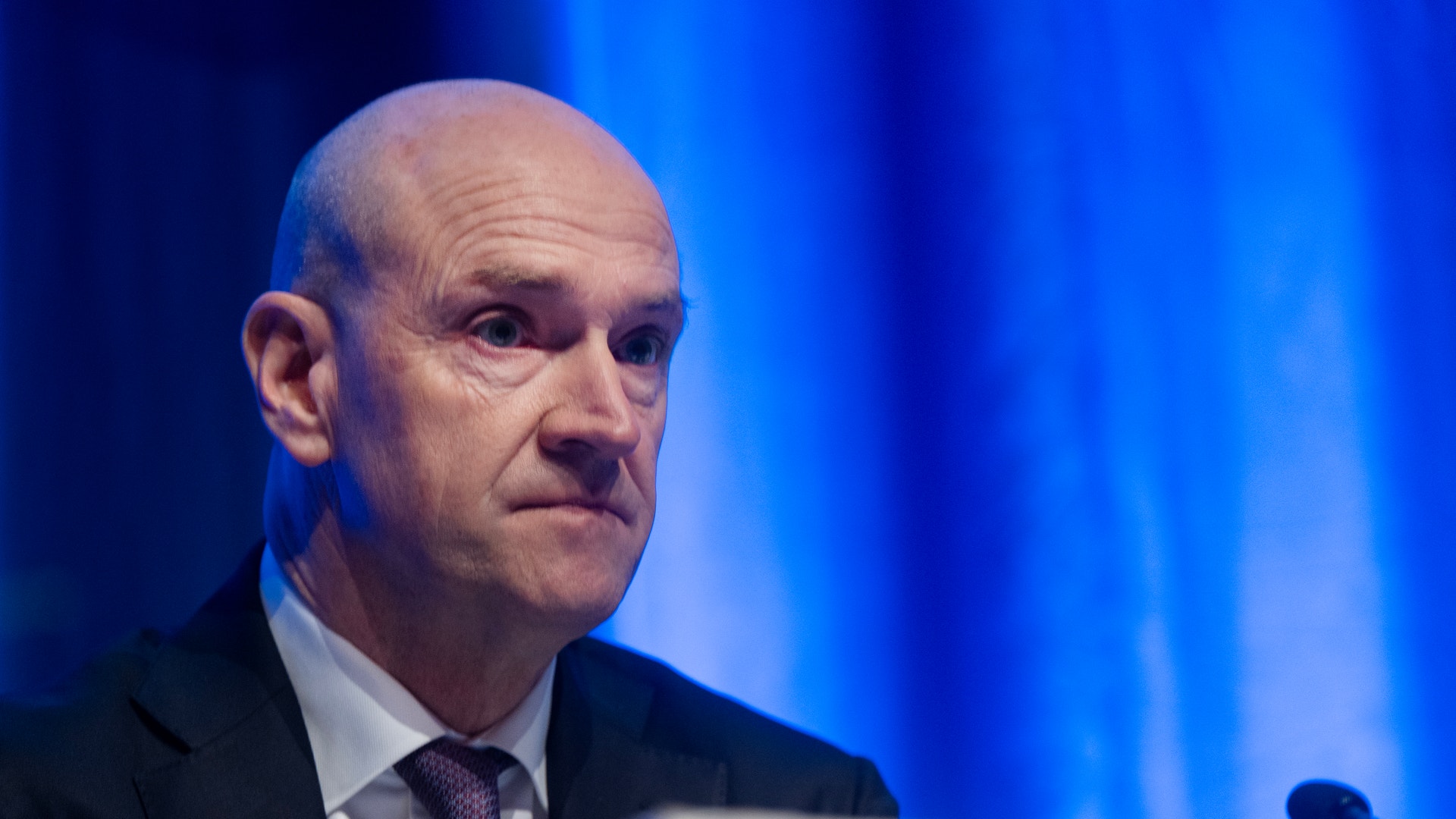 En man i kostym och slips talar vid ett podium mot en blå bakgrund.