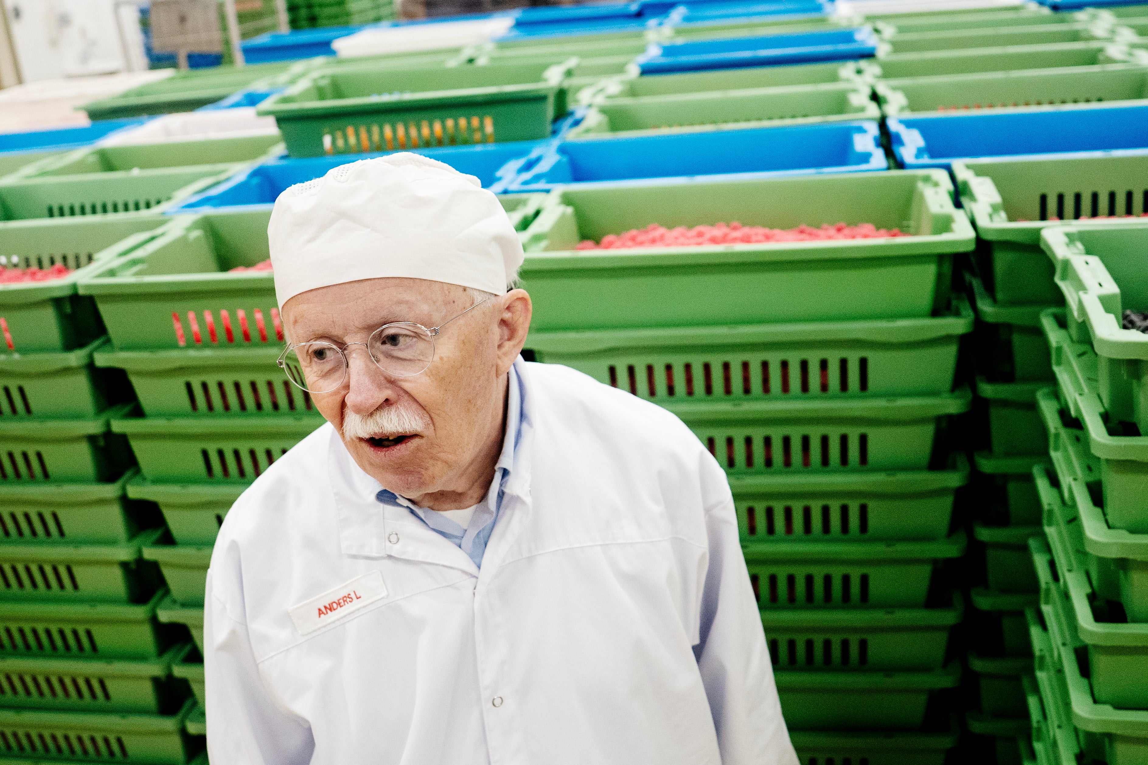 En äldre man i vita arbetskläder och huvudduk står framför stora gröna och blå plastlådor med livsmedel i en produktionsmiljö.