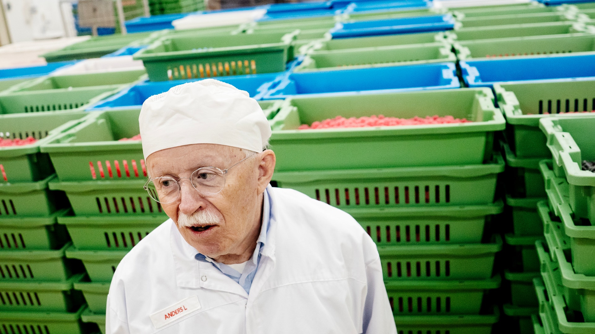 En äldre man i vita arbetskläder och huvudduk står framför stora gröna och blå plastlådor med livsmedel i en produktionsmiljö.
