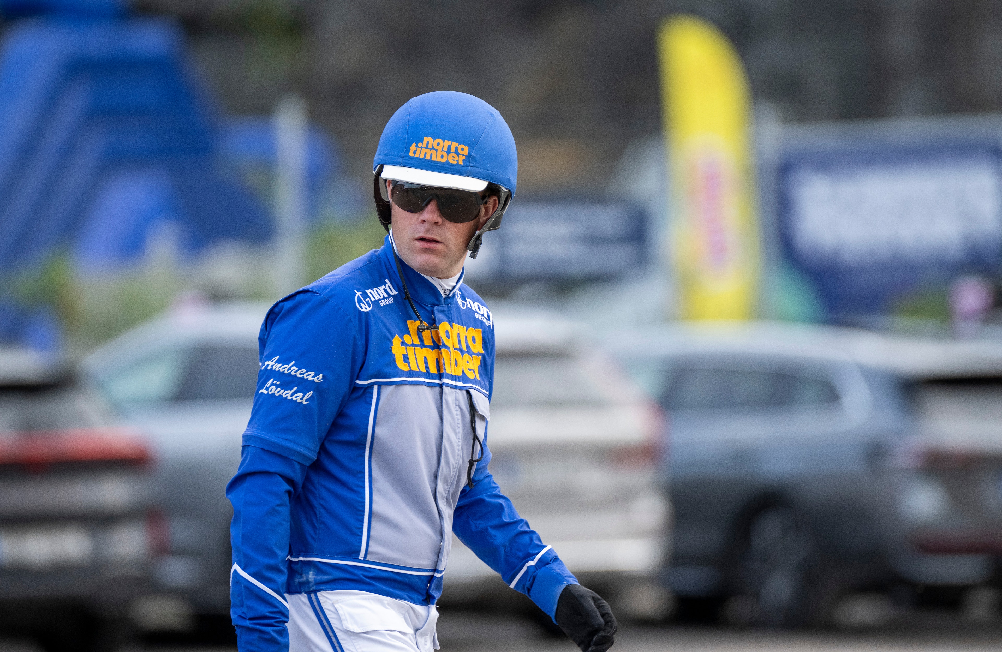 Cyklist i blå och vit racingdräkt med hjälm och solglasögon står framför parkerade bilar.