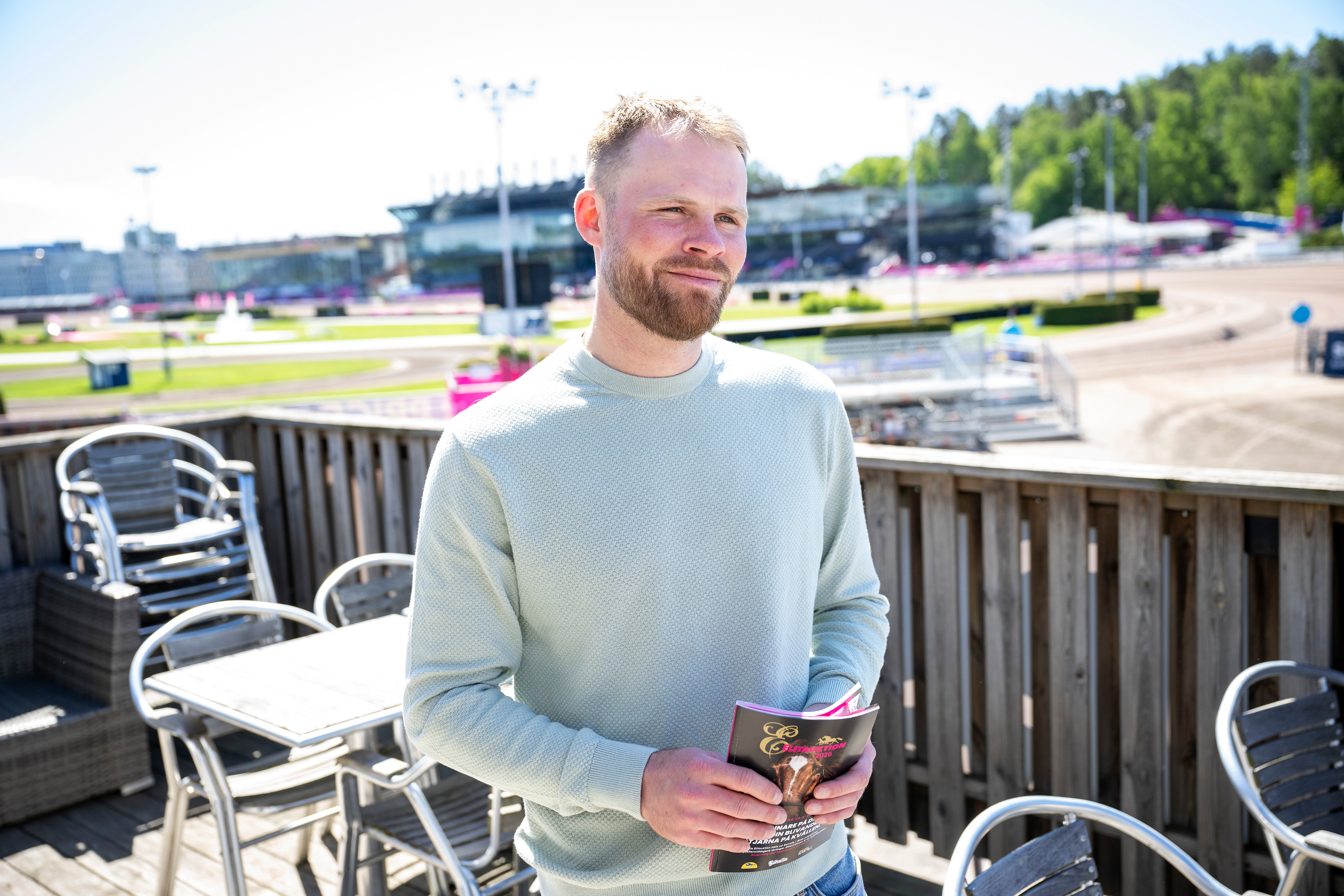 En man står på en terrass med utsikt över Solvalla travbana. Han är klädd i en ljusblå tröja och håller i en dryck.