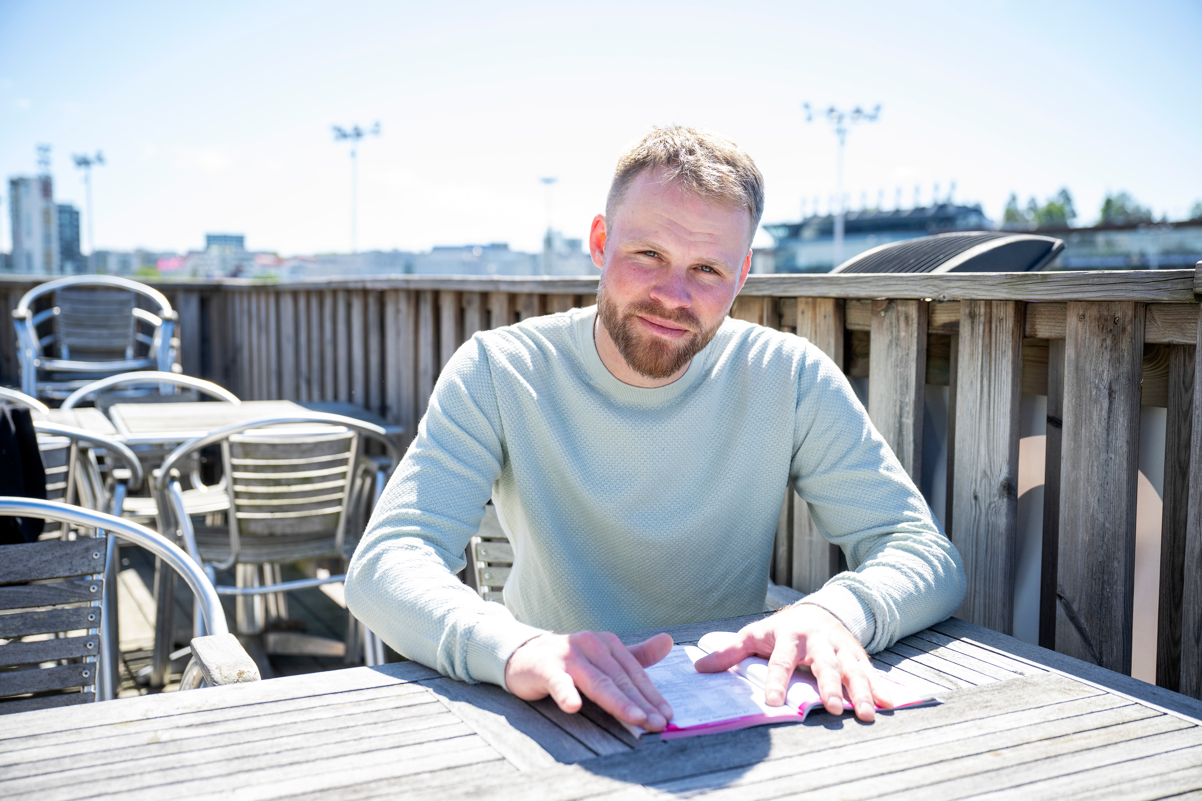 En man i ljusblå tröja sitter vid ett bord på en terrass med stadsvy i bakgrunden.