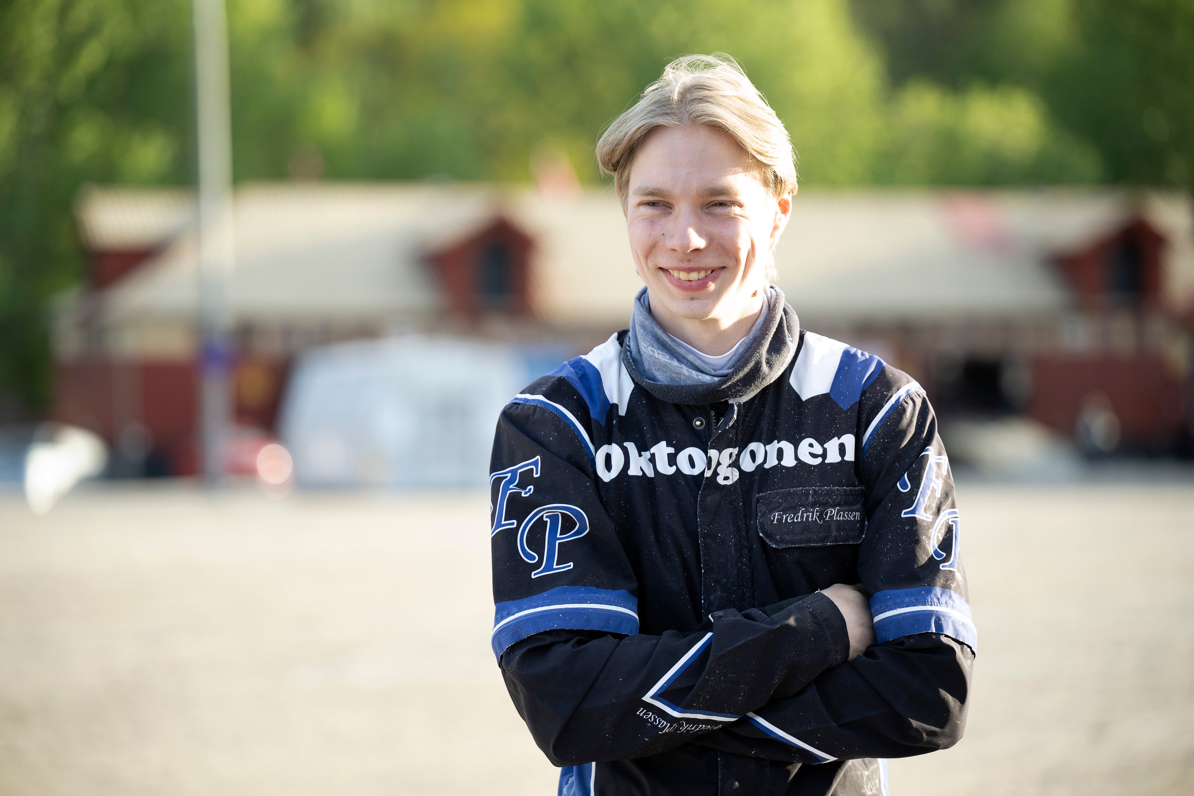 Porträtt av en ung person i blå och svart jacka på en hästhall. Personen står framför stall och leende mot kameran.