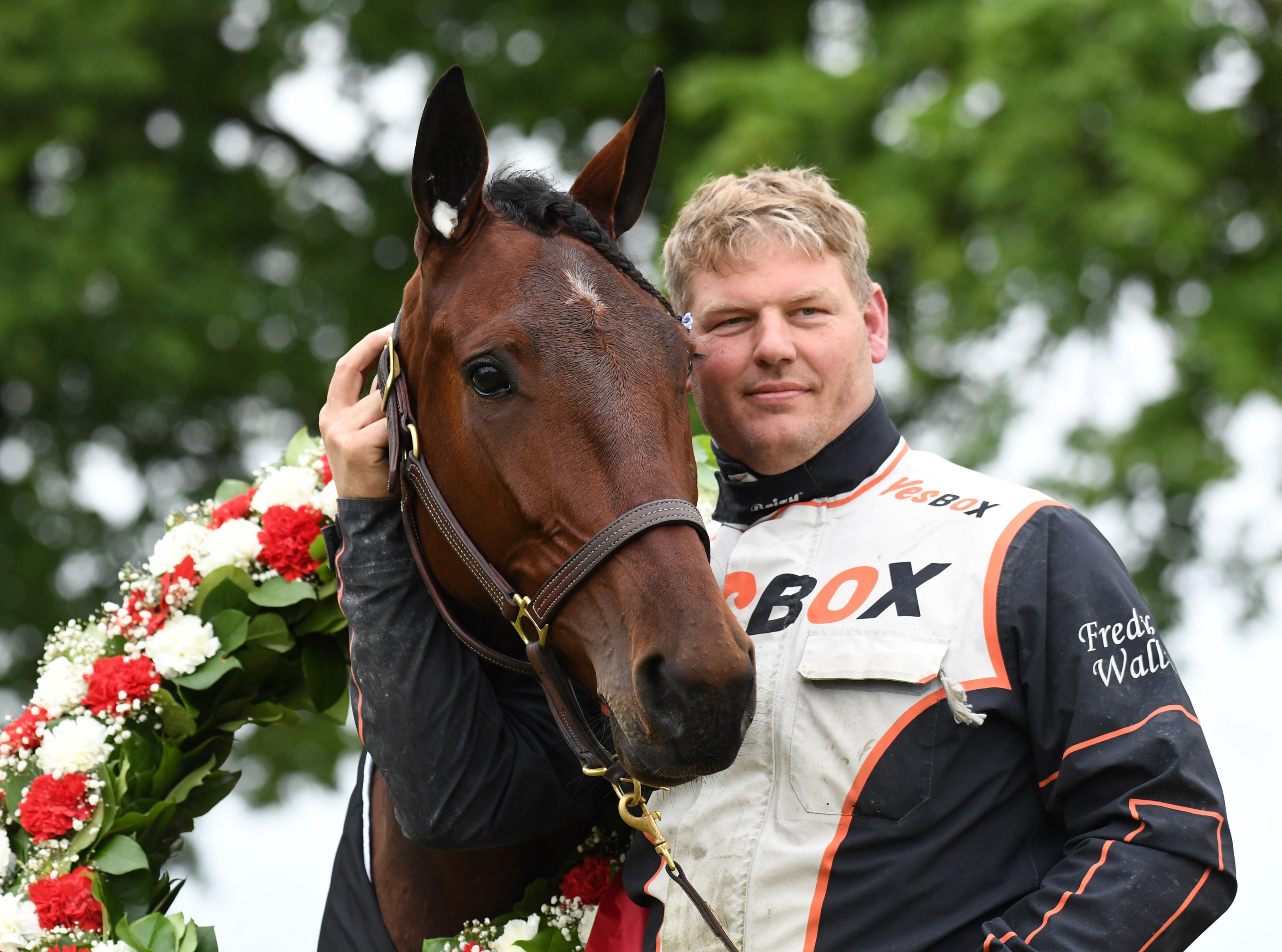 En man i racing-overall står tillsammans med en brun häst som bär en blomsterkrans runt halsen. Mannen håller hästen och ler mot kameran. Grön löv i bakgrunden.