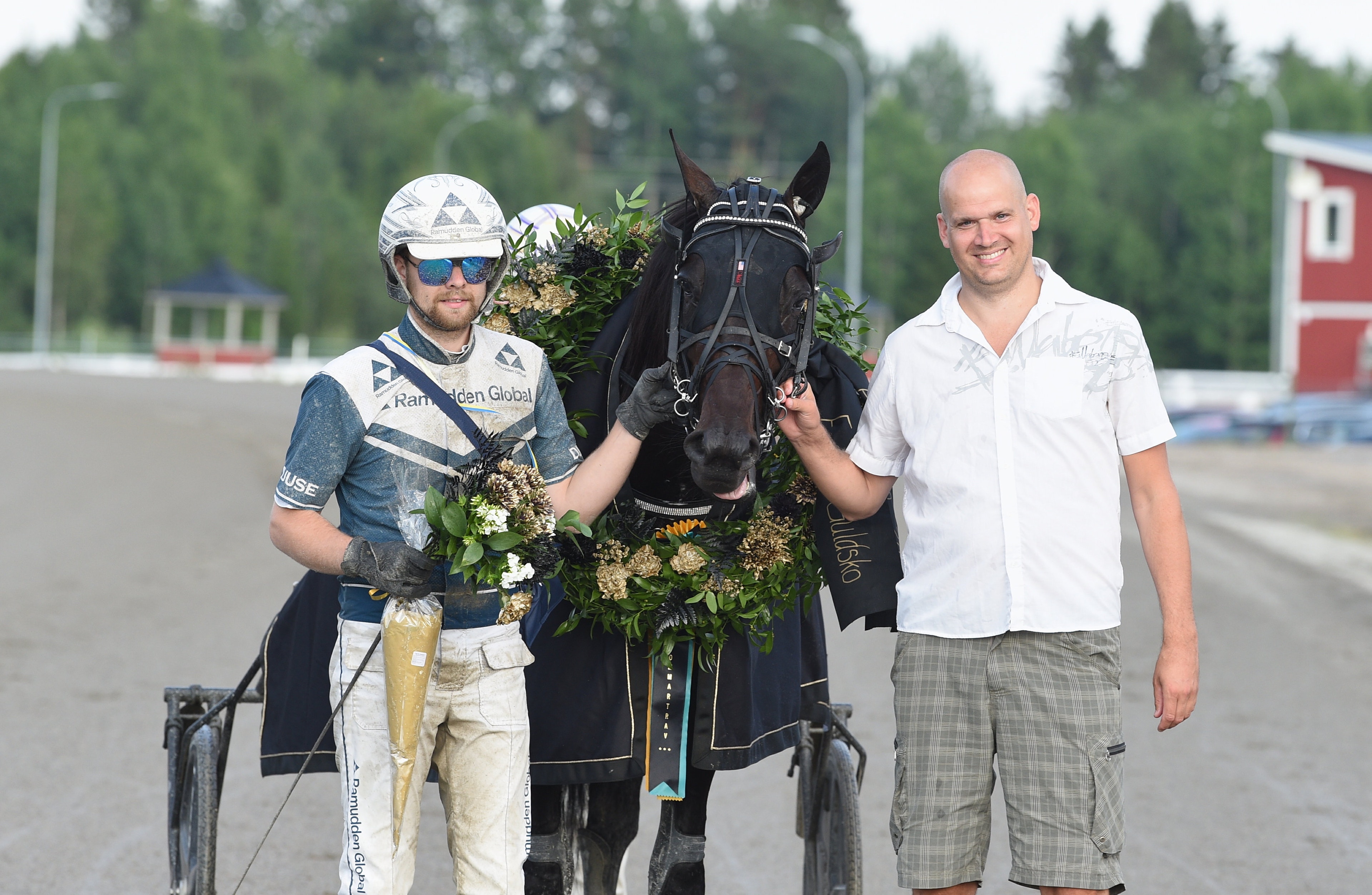 En kusks och en man poserar med en svart travhäst som bär en vinnarkrans efter ett lopp.