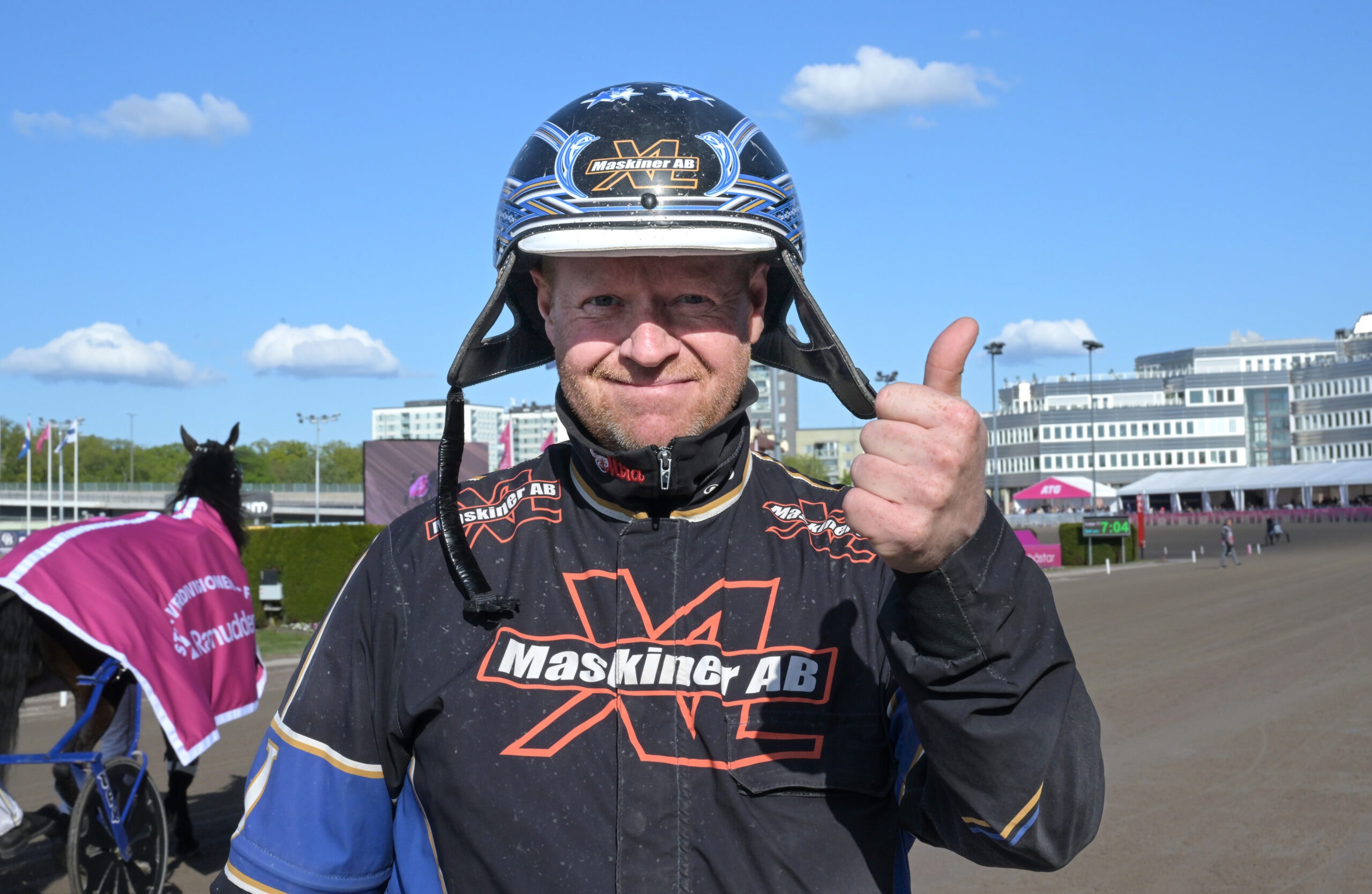 Man i svart racing-dräkt och motorsport-hjälm som ler och gör tummen upp-gesten. I bakgrunden syns en öppen plats med byggnader.
