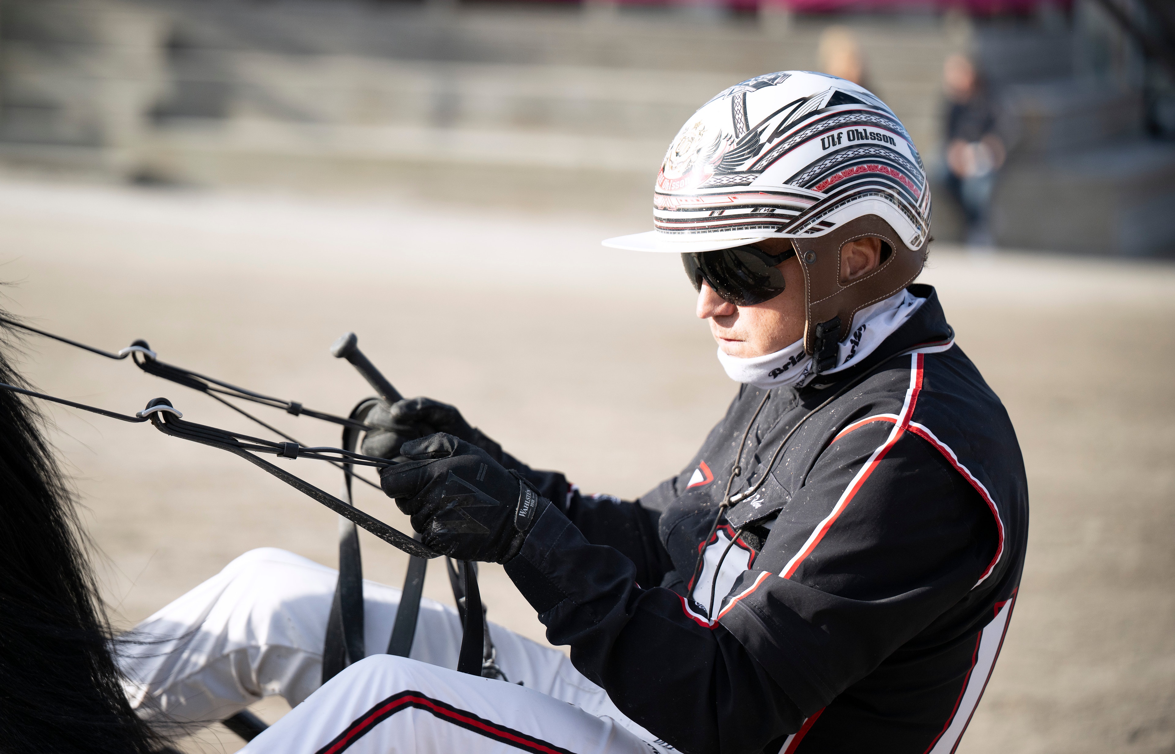 Kusksportare i vit hjälm och svart tävlingsdräkt håller i tyglar under ett travlopp på Solvalla.
