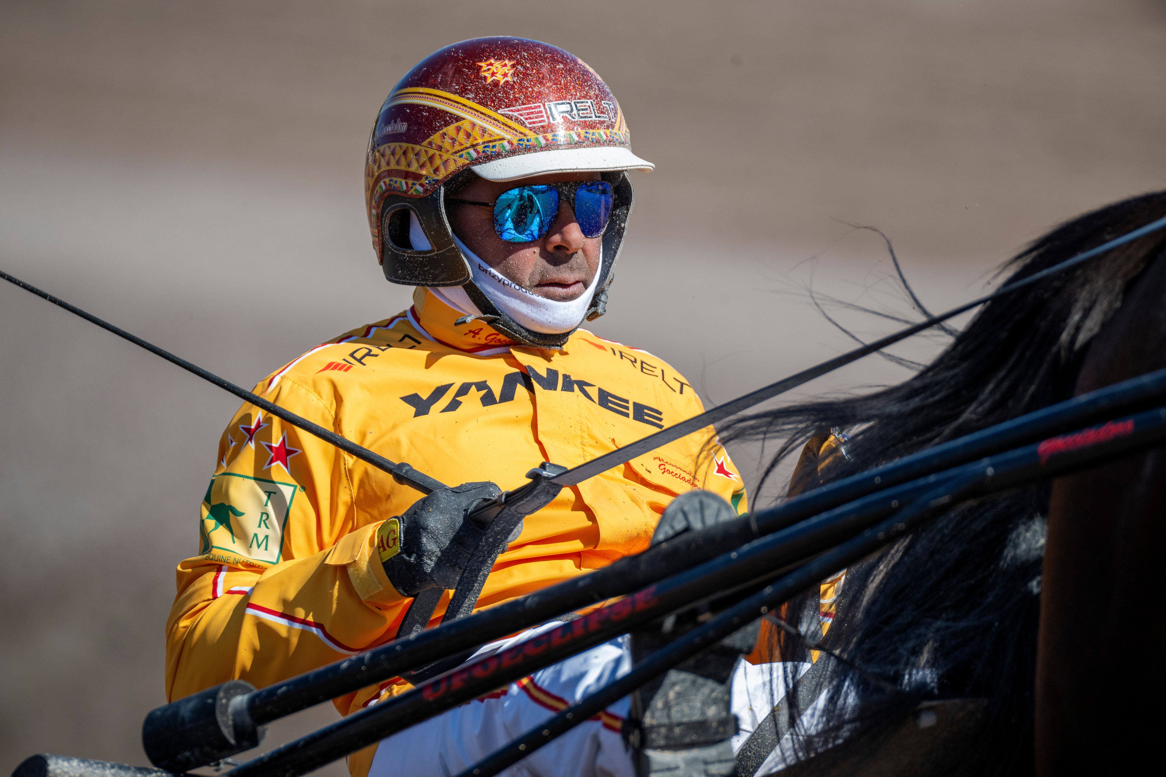 En jockey i gul tävlingsdräkt och röd hjälm med blå glasögon sitter på en häst under tävling.