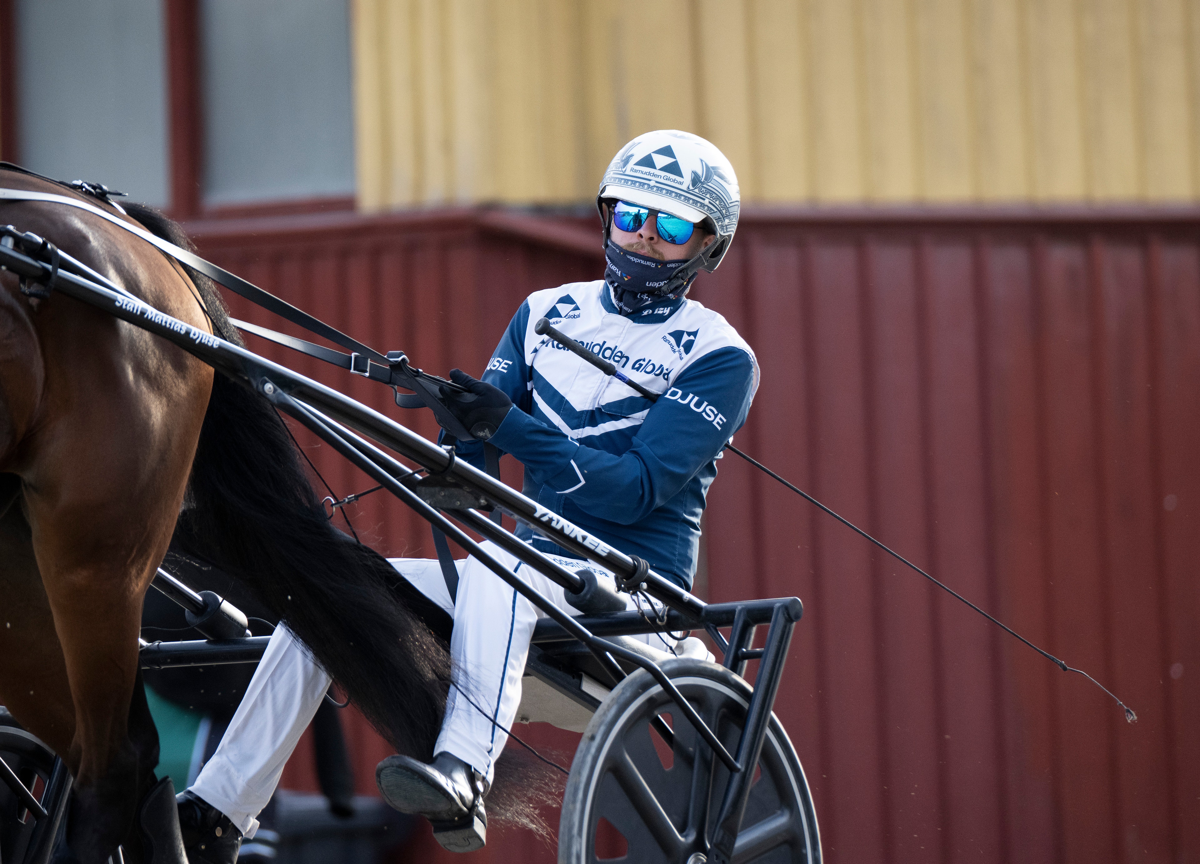 Kusk i blå och vit tävlingsdräkt sitter i kuskvagn bakom en brun travhäst. Kusken bär hjälm och solglasögon. I bakgrunden syns en rödbrun stallbyggnad.