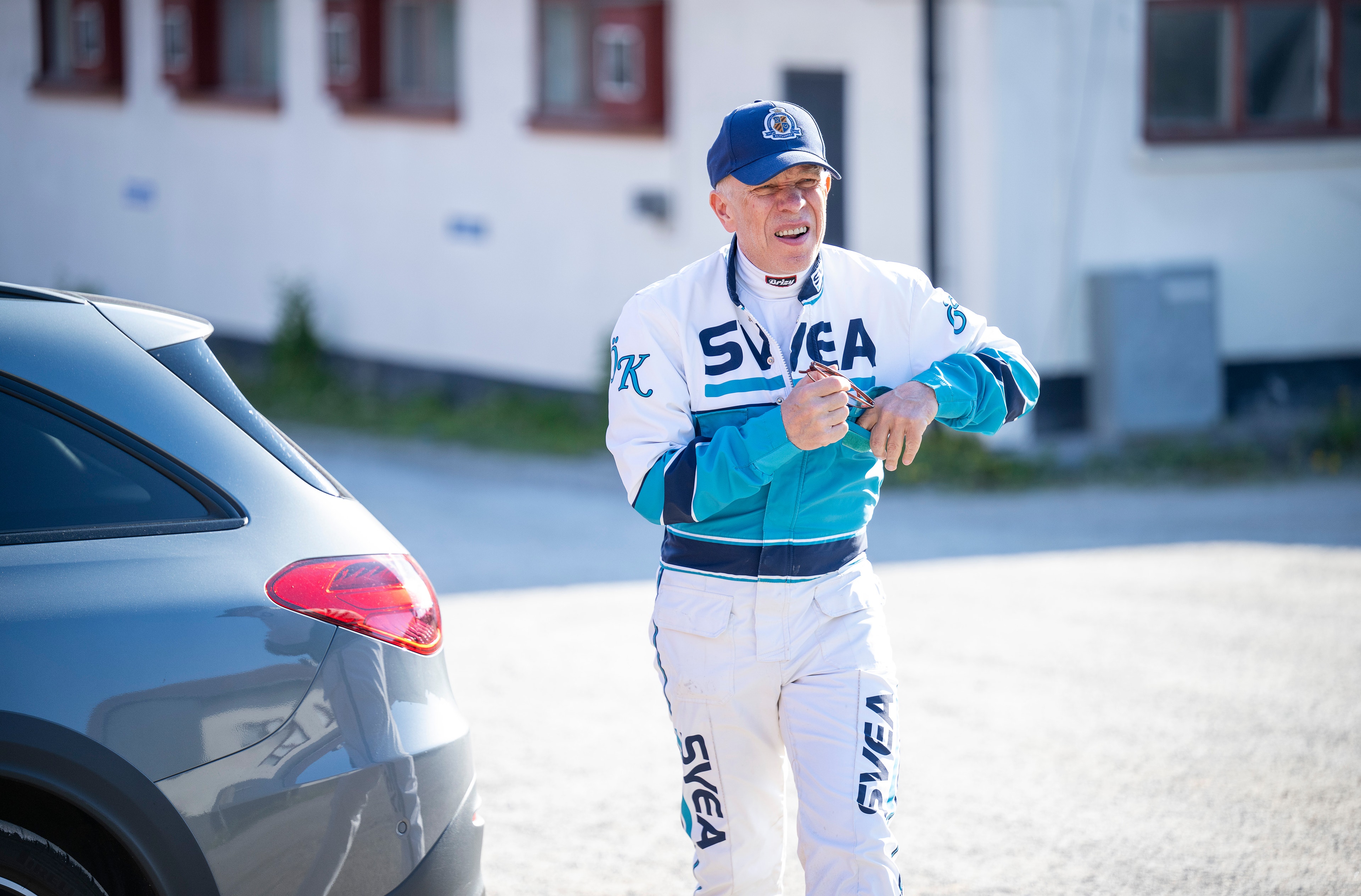 En man i vit och turkos träningskläder med texten SVEA står bredvid en grå bil på en parkeringsplats. Han bär en blå keps.