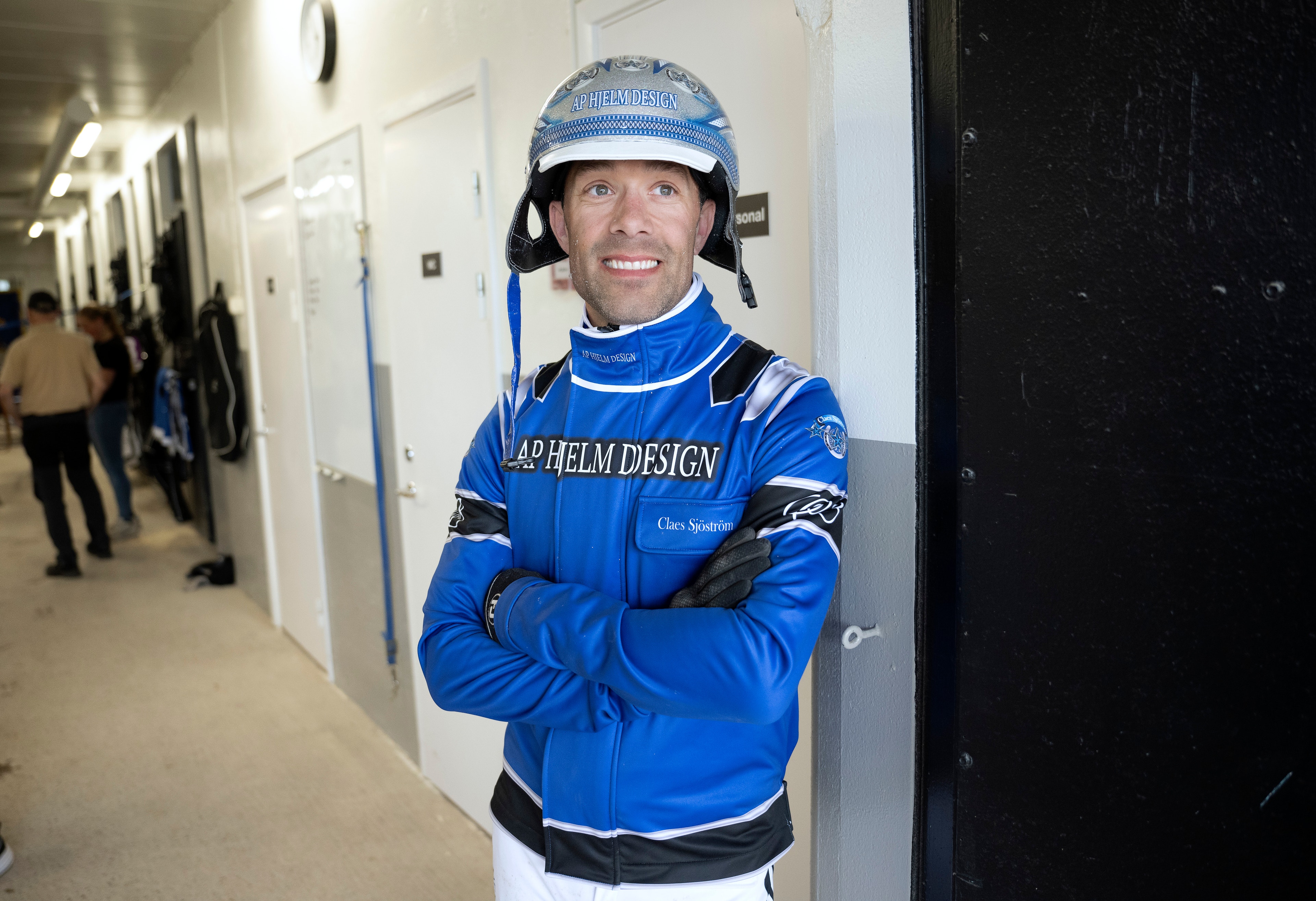 Racerförare i blå racing-overall och hjälm står med armarna i kors och poserar för kameran i en korridor.