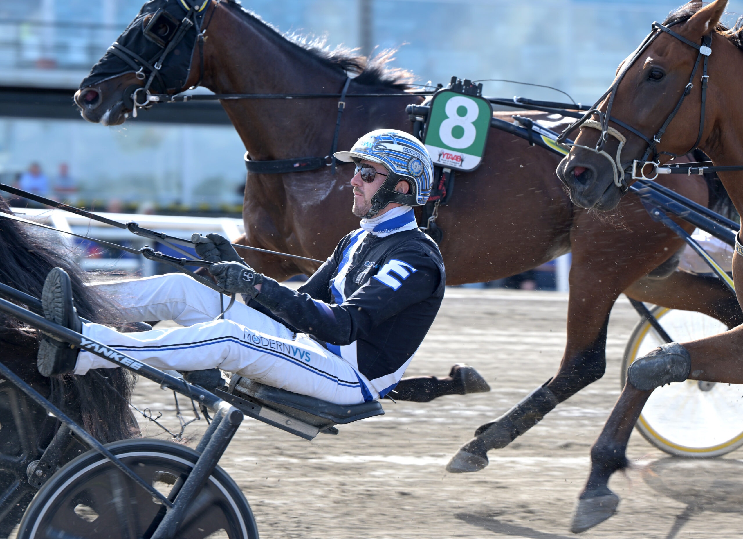 En travkusk i sulky kör i högt tempo under ett travlopp, med hästar i galopp runt omkring.