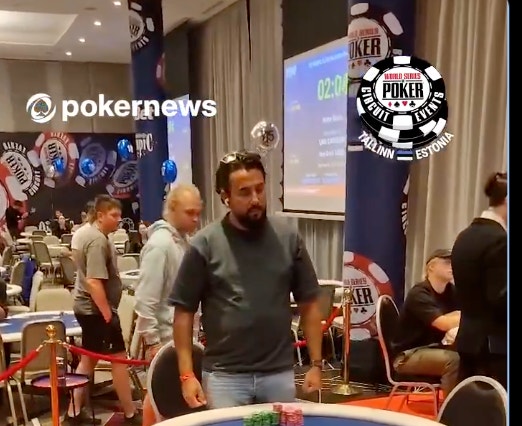 Interiör från ett pokerevent med deltagare vid pokerbord. I bakgrunden syns pokernews-logotyp och World Series of Poker-banderoller. Flera spelare är samlade vid ett rött pokerbord med pokermarker, medan andra står omkring. Stora skärmar och pokersymboler är synliga på väggarna.