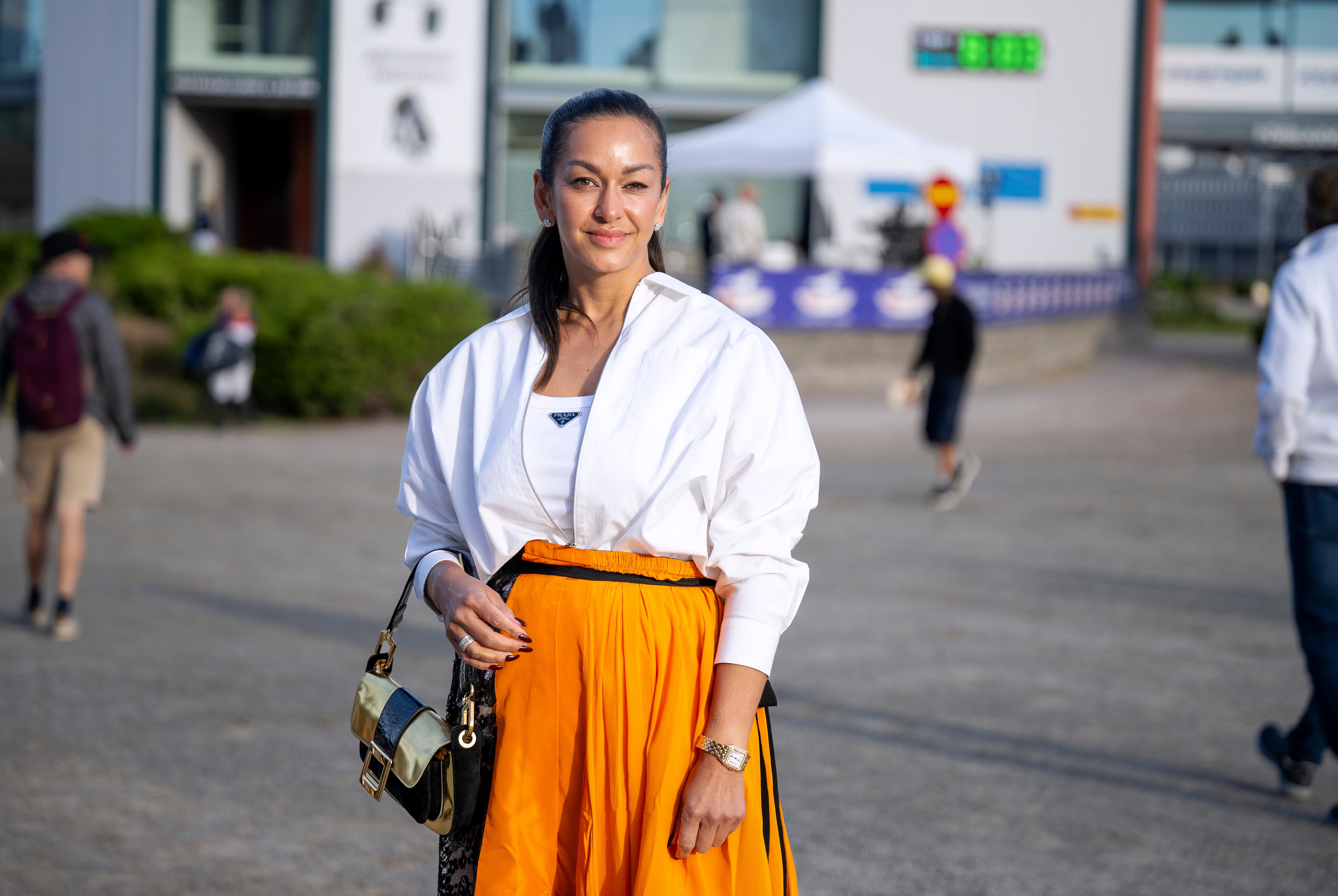 Porträtt av en kvinna som står utomhus på en öppen plats. Hon är klädd i vit skjorta och orange kjol, och bär en väska över axeln.
