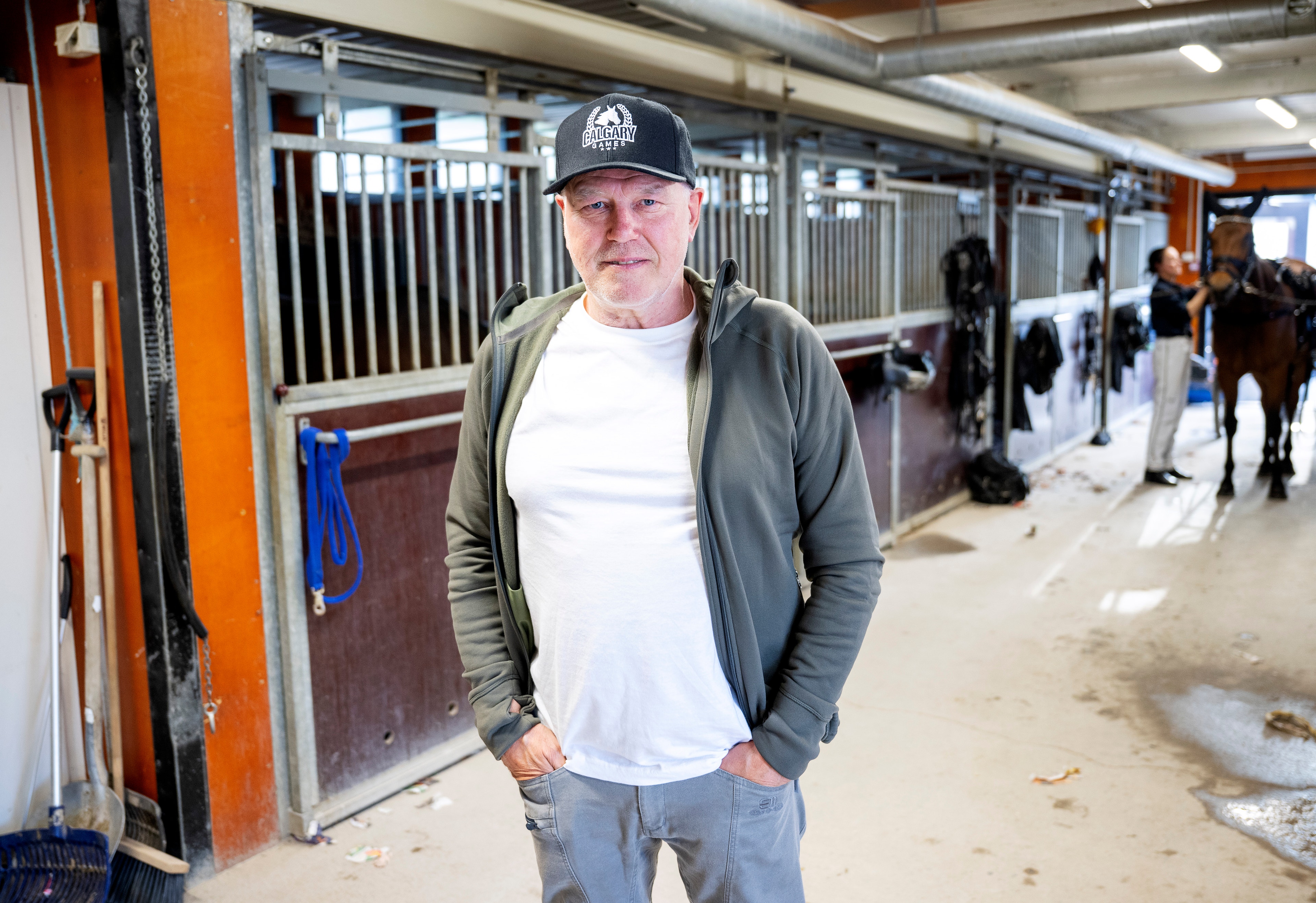 En man i mössa och grå jacka står i en häststall. Han är omgiven av hästboxar och andra människor som arbetar med hästarna i bakgrunden.