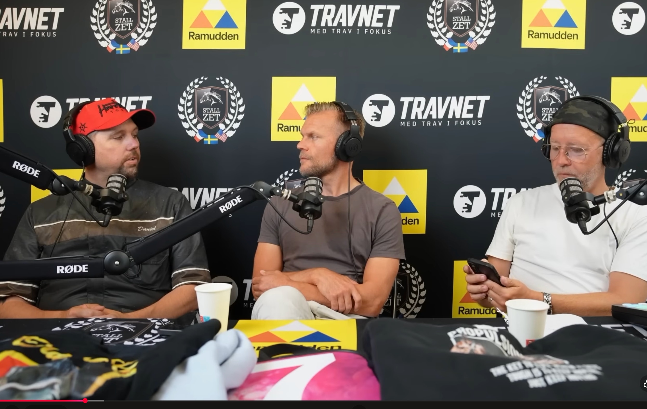 Tre män sitter vid en podcastinspelning med mikrofoner framför en sponsorvägg med logotyper för Travnet och Ramudden.