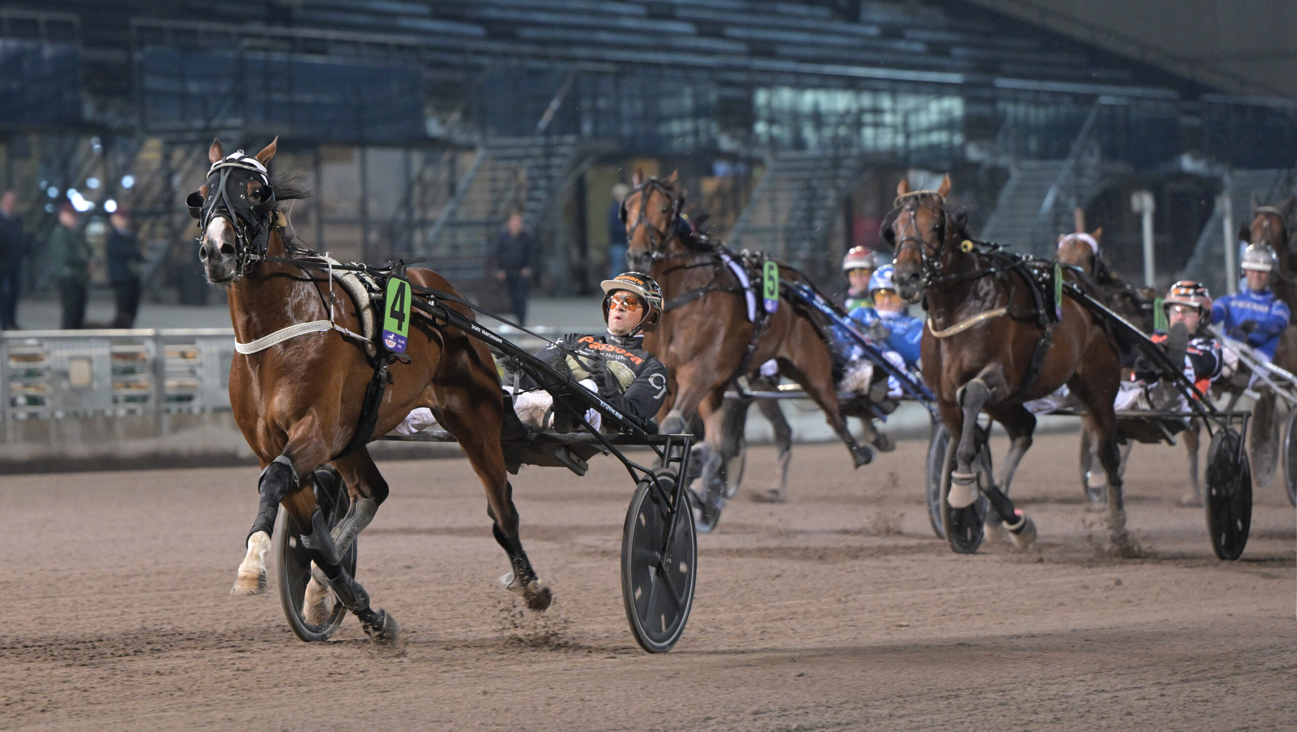 Travhästar med kuskar i sulkyn under ett travlopp på en sandbana. Hästen längst fram leder loppet, med flera hästar tätt bakom sig och en läktare i bakgrunden.