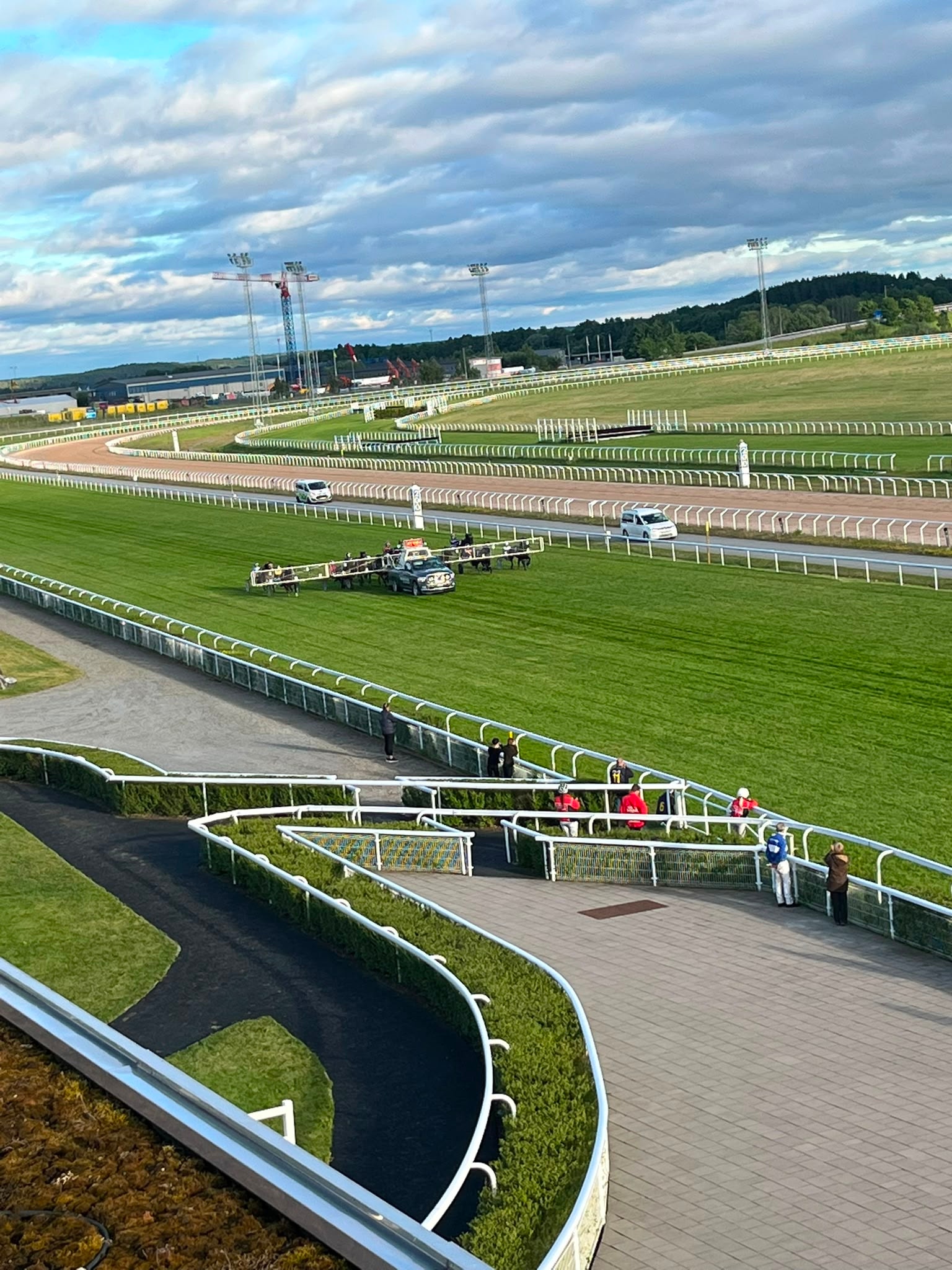 Luftfoto över en modern travbana med gröna racebanan, vita räcken och publik på en utomhusplattform. Blå himmel med moln och industrianläggningar i bakgrunden.