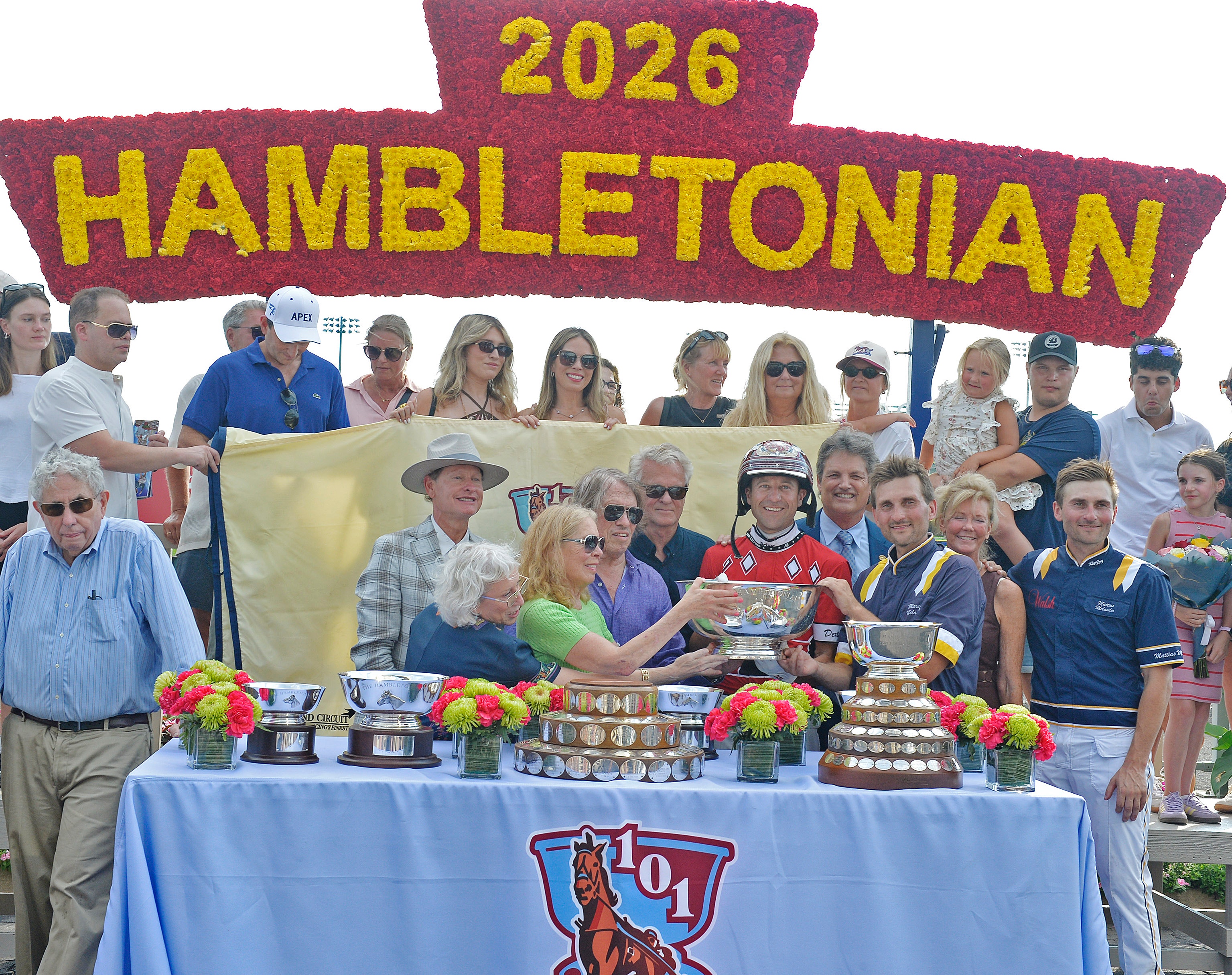 Grupp av människor samlade vid ett prisbord under en banderoll för Hambletonian 2026-tävlingen. Flera pokaler och troféer är utställda på bordet tillsammans med blommor.