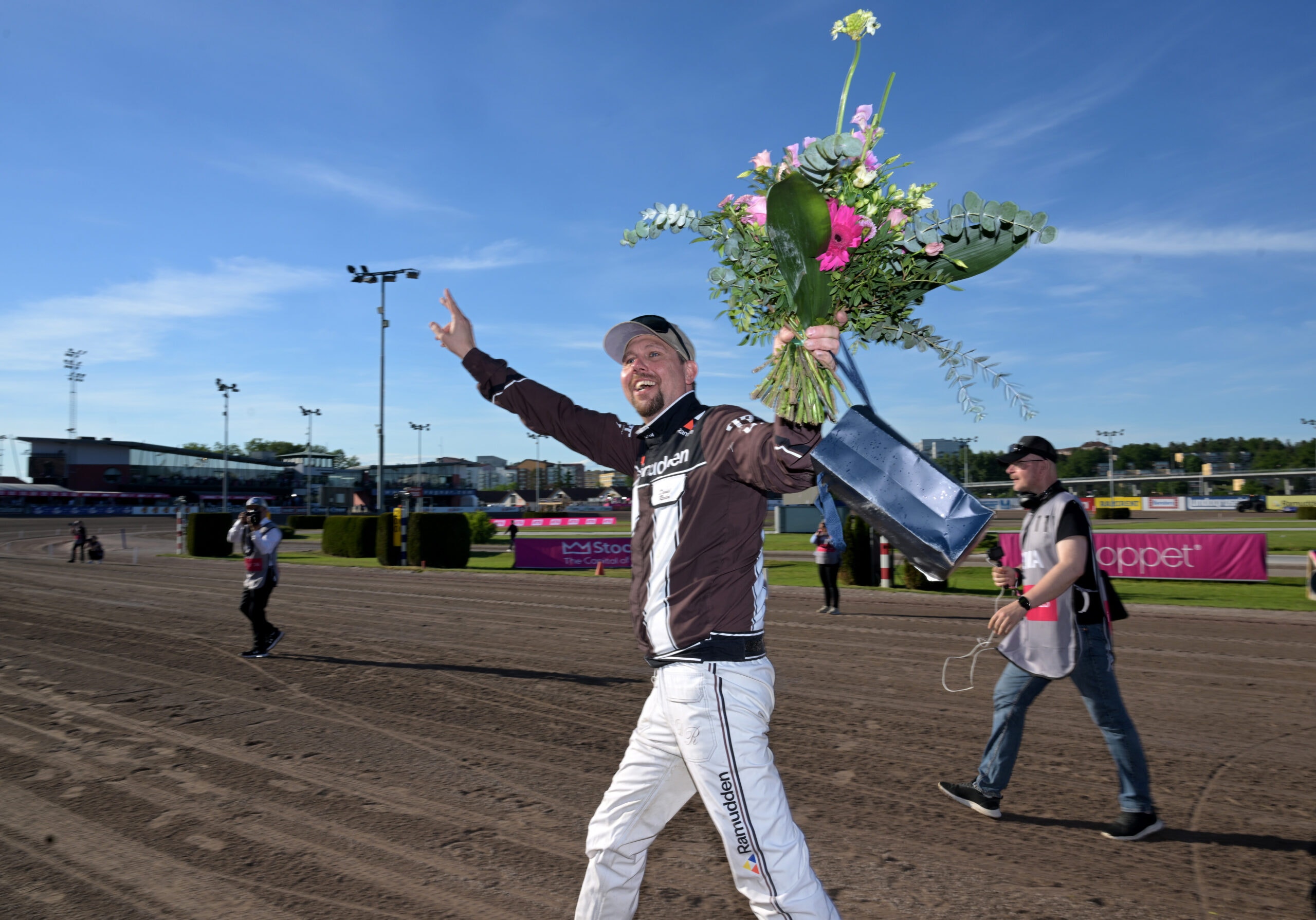 En jockey firar en seger på travbanan och håller upp en blomsterbukett med en glad gest.